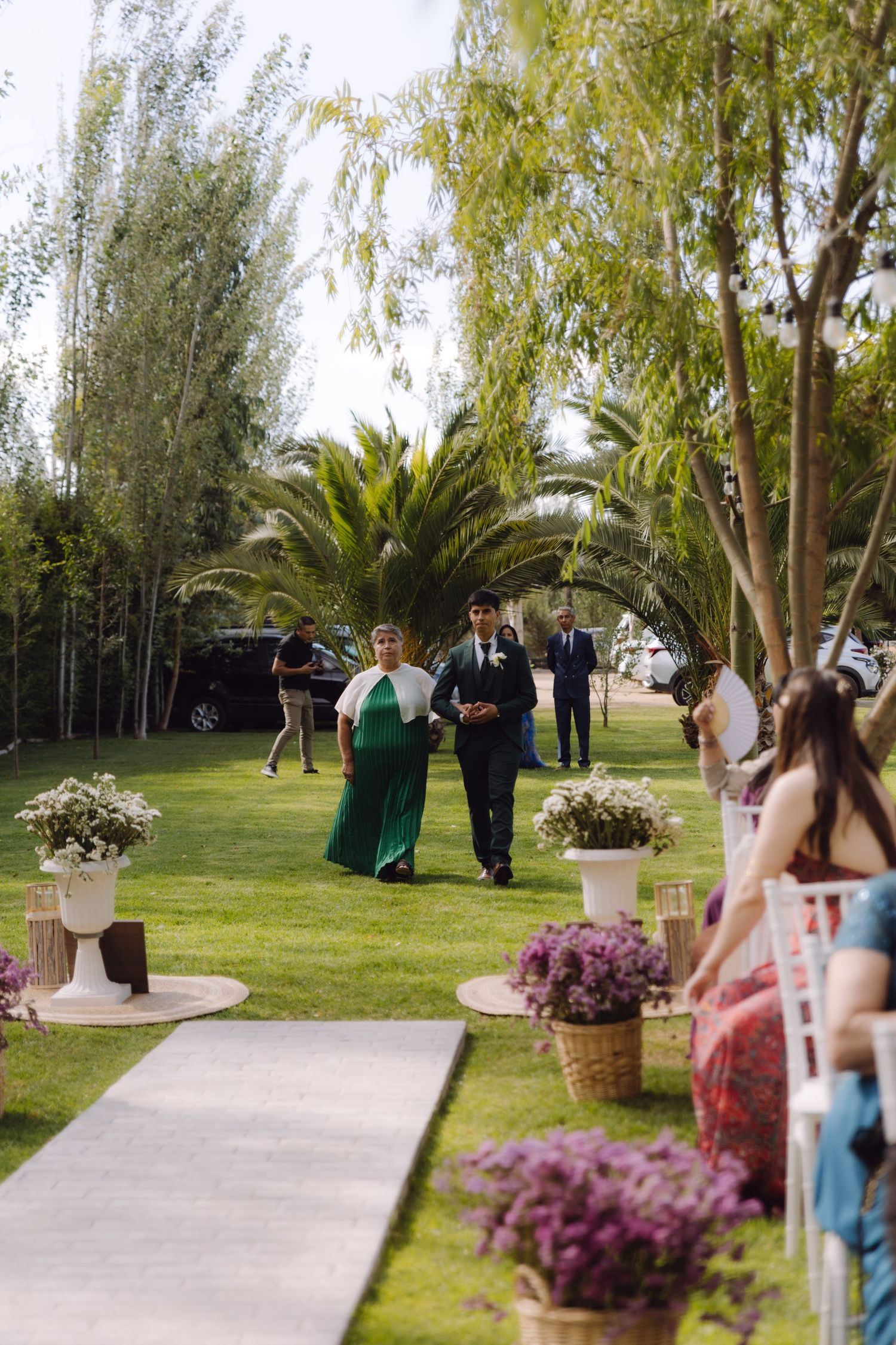 Groom escorted by mother down outdoor wedding aisle lined with flower arrangements.