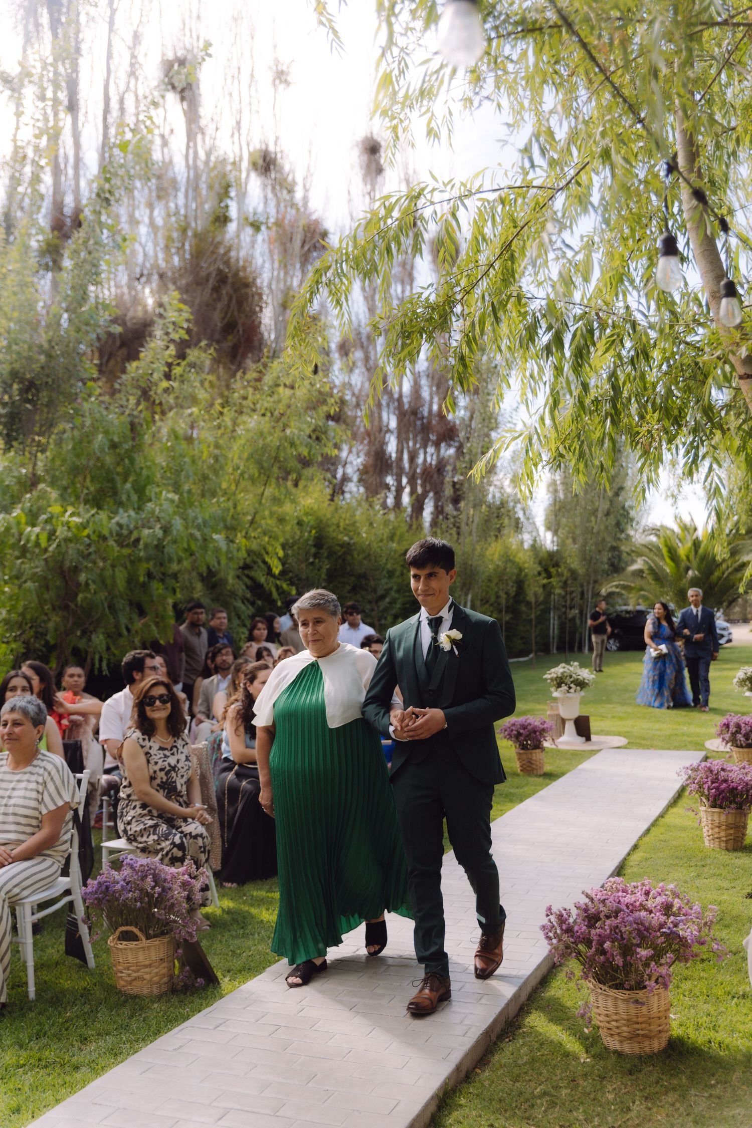 Groom walking mother down garden wedding aisle with seated guests watching ceremony.