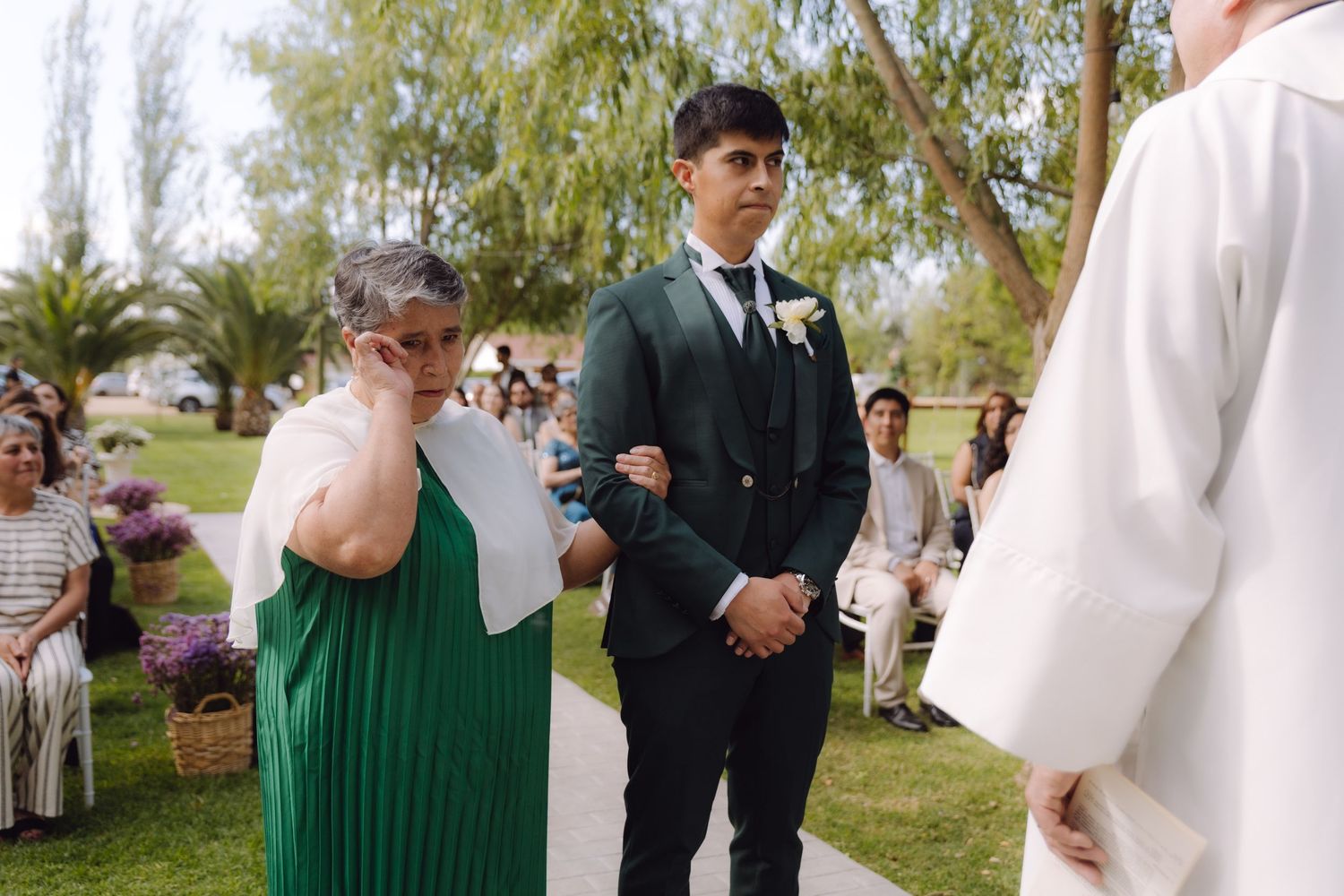 Emotional mother in green dress wipes tears beside groom in dark green suit at outdoor wedding ceremony.