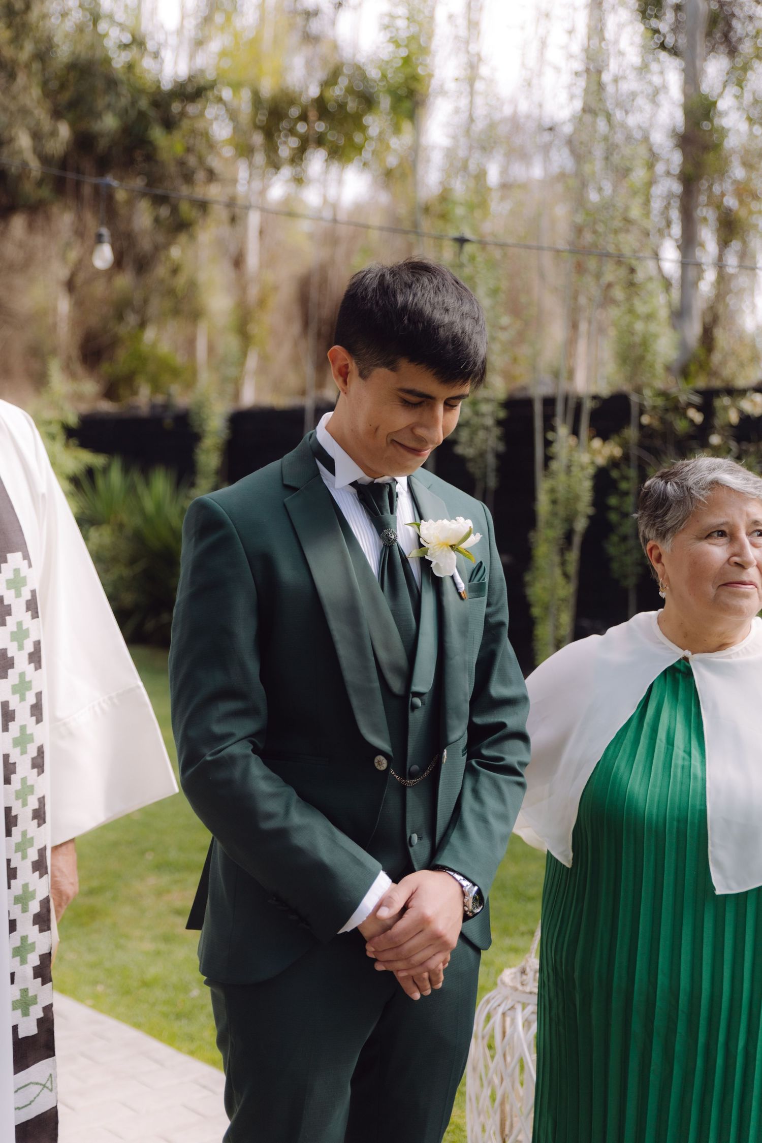 Handsome groom in dark green three-piece suit with white boutonniere stands at outdoor wedding altar.