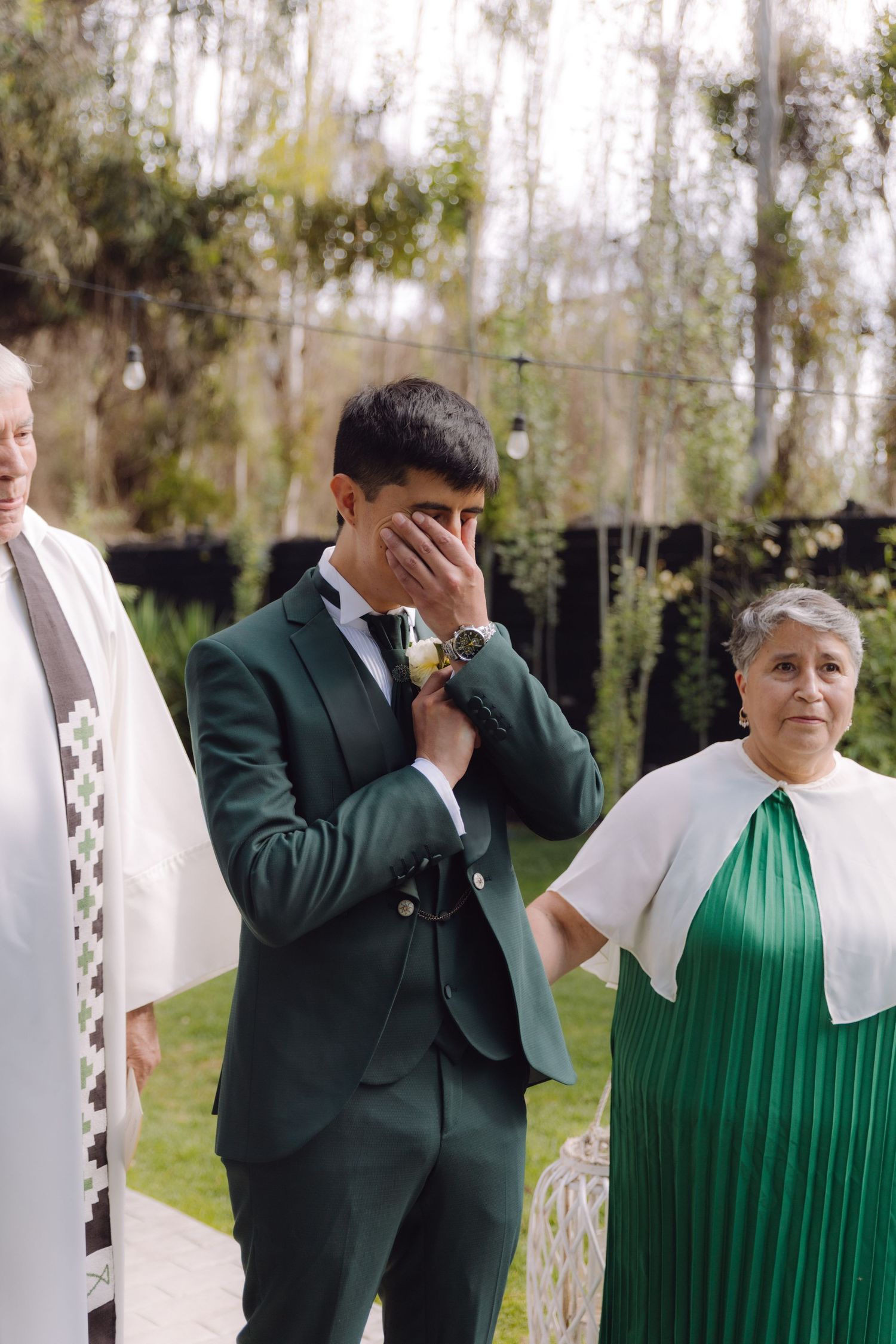 Emotional groom in green suit covers face with hand during touching outdoor wedding ceremony moment.
