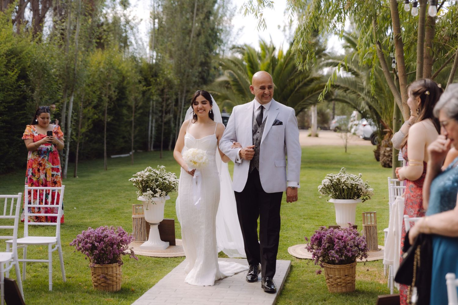 Bride in white gown with bouquet walks down aisle with escort at elegant outdoor garden wedding.