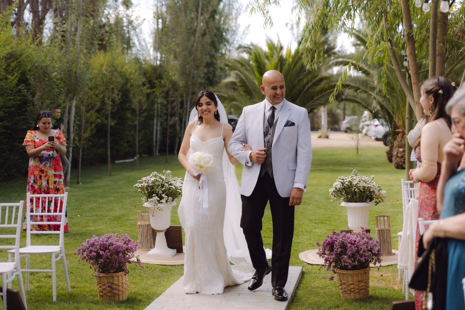 Elegant bride in fitted white dress and veil walks down flower-lined aisle with her escort.