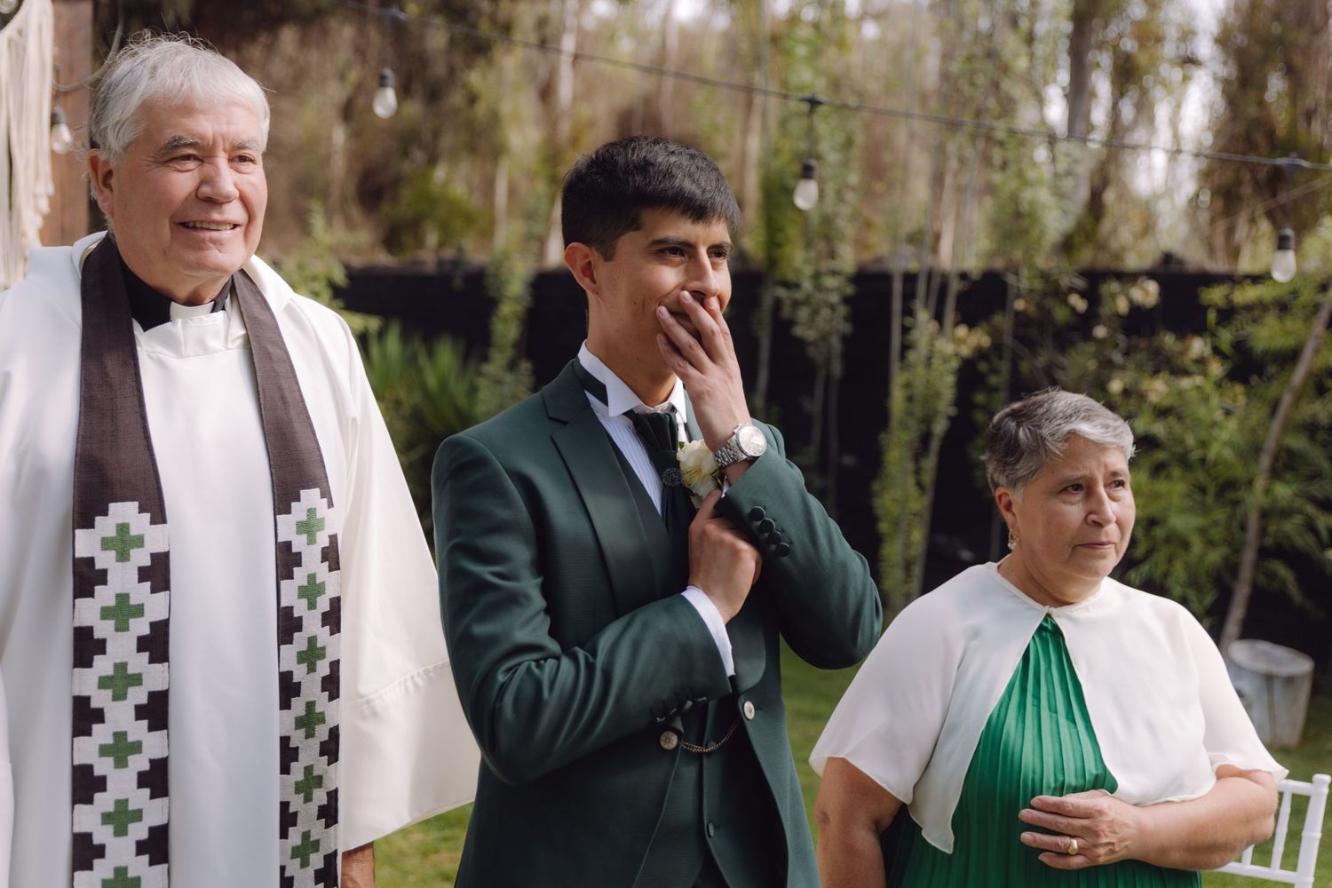 Groom in green suit covers mouth emotionally beside officiant and older woman at outdoor ceremony.
