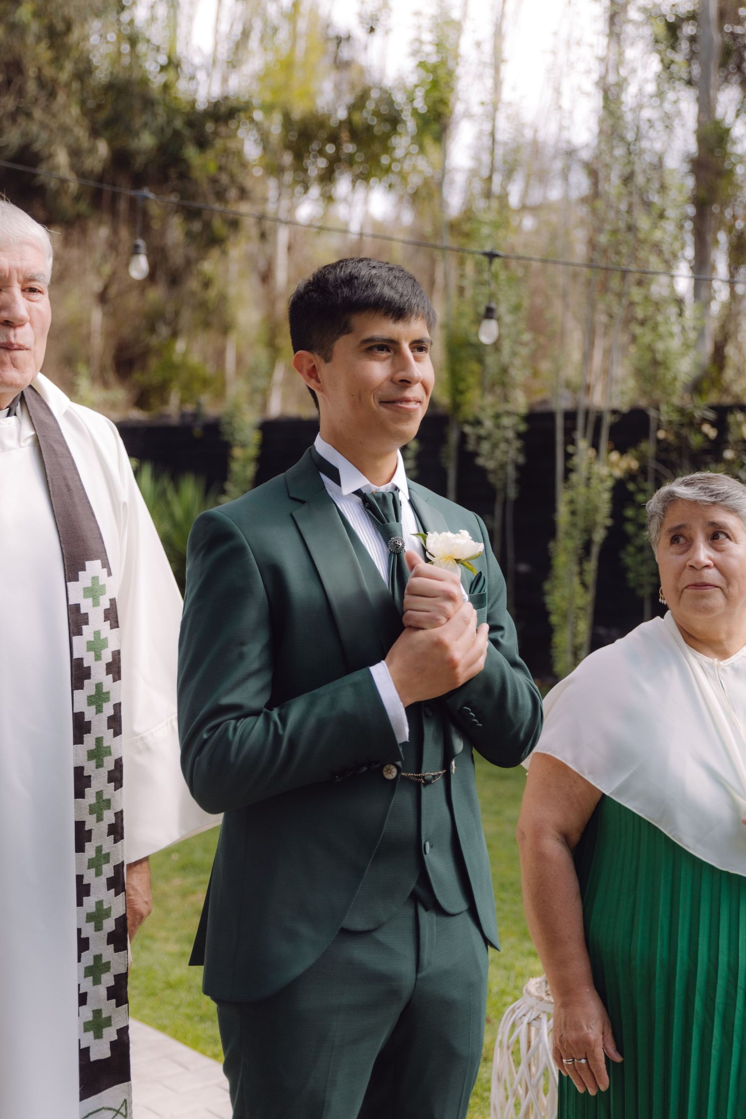 Dapper groom in dark green suit clasps hands nervously at outdoor garden wedding ceremony.
