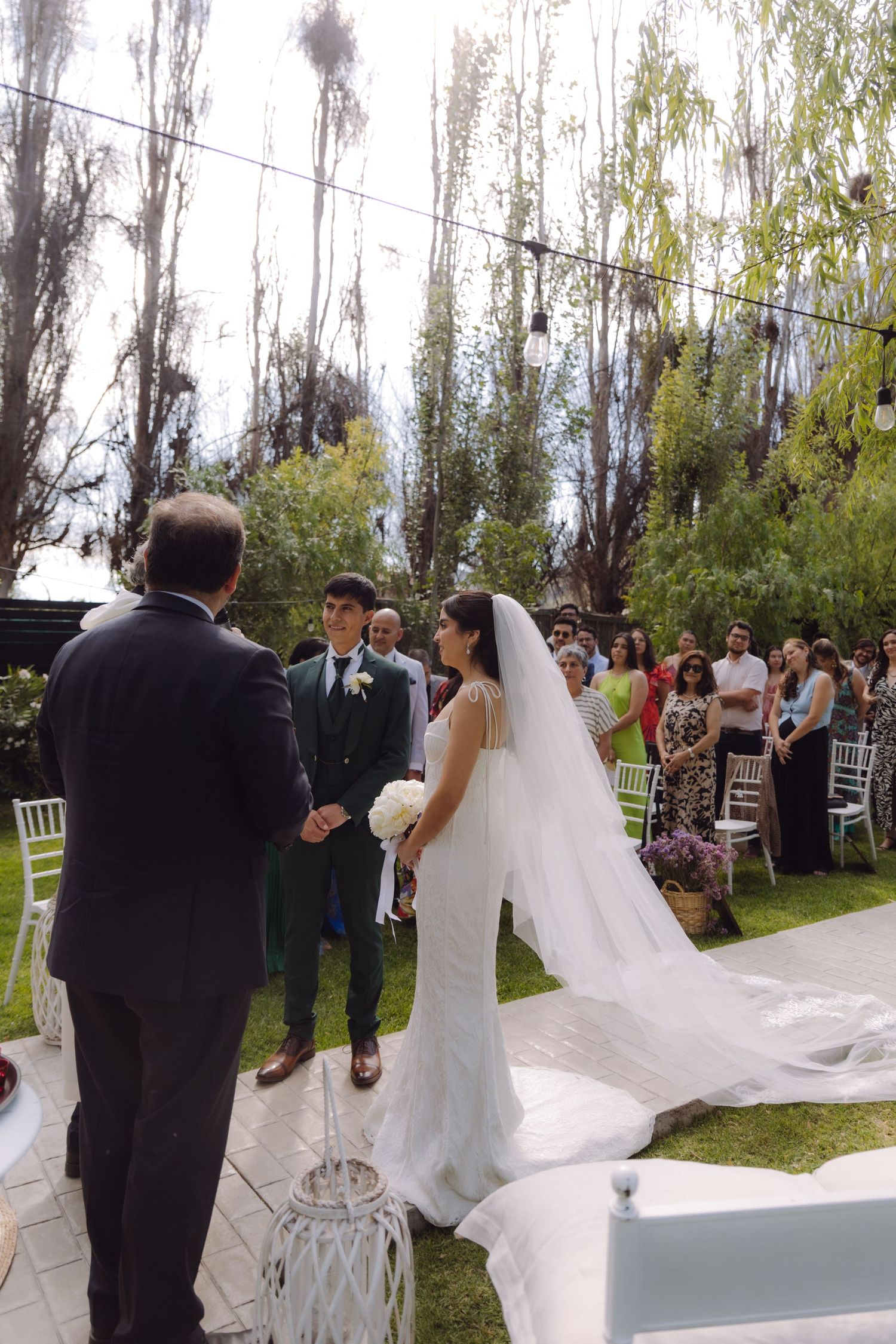 Bride in long veil and white gown faces groom at romantic outdoor garden wedding ceremony.