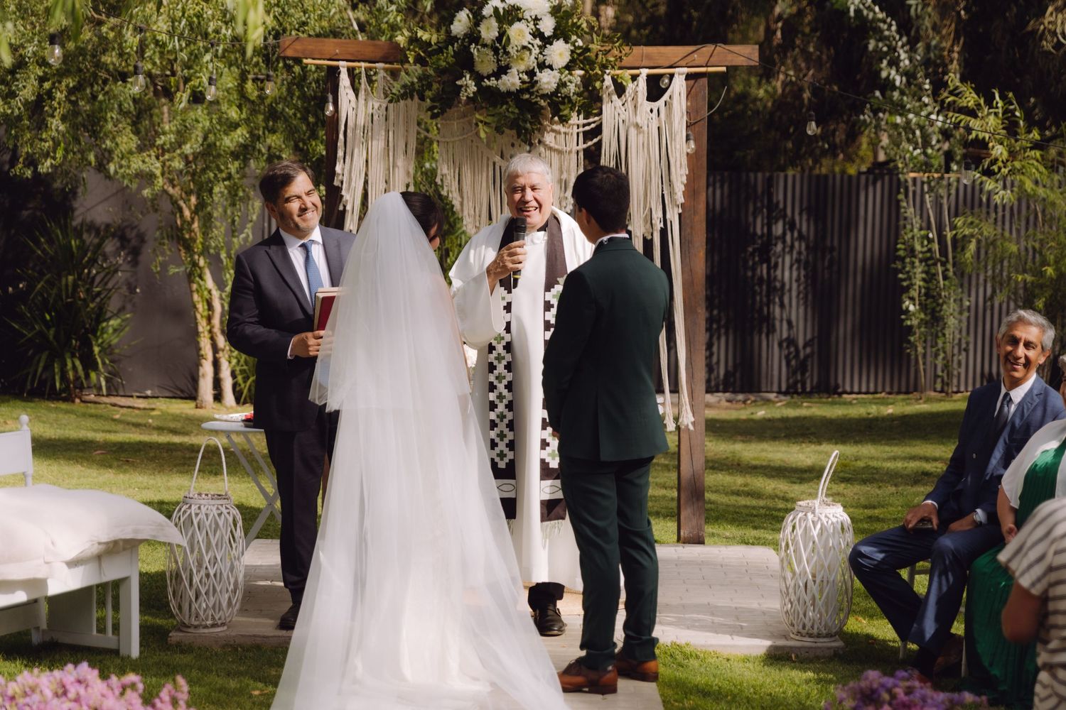 Wedding officiant speaks beneath floral macrame arch as bride in long veil stands at altar.