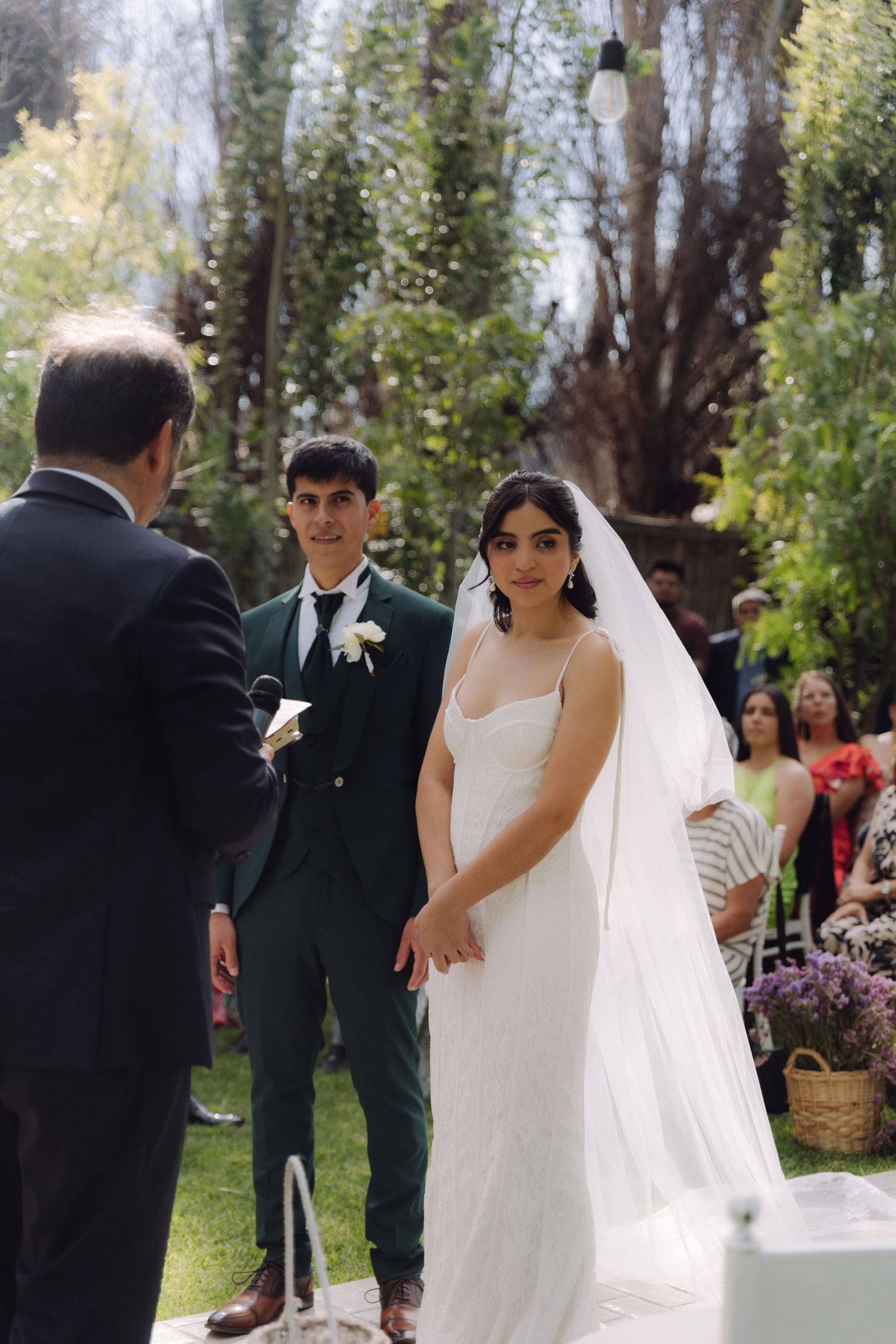 Bride in white gown and veil stands at outdoor wedding altar with groom in dark green suit.
