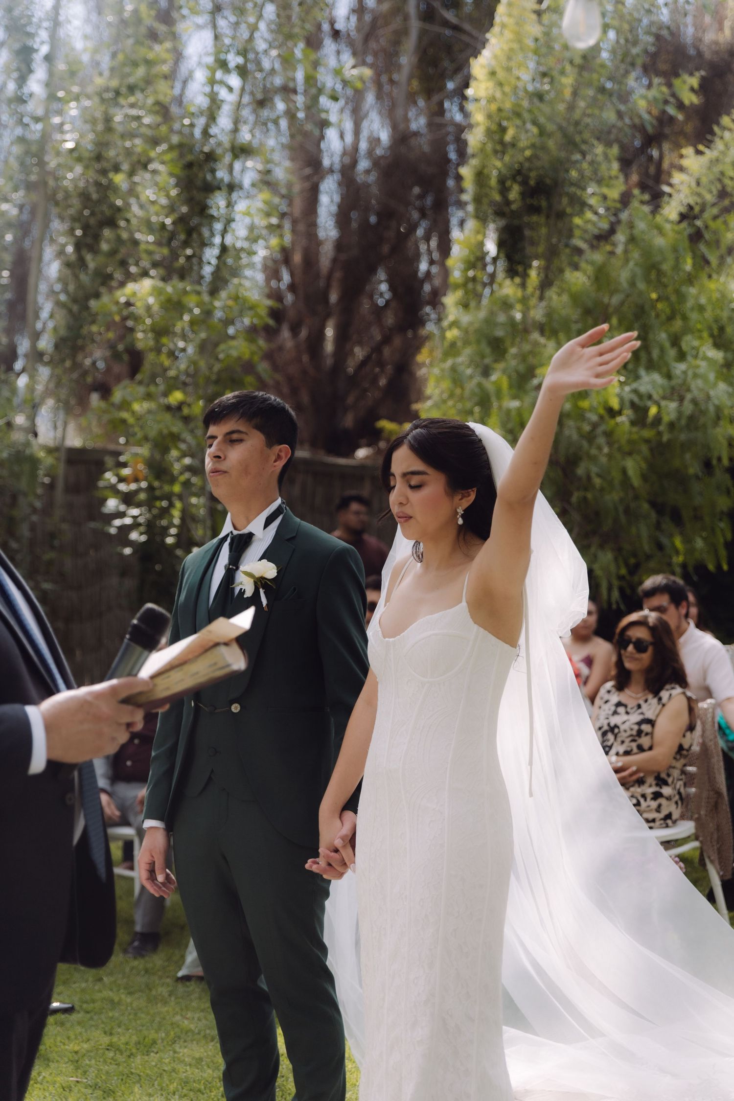 Joyful bride raises her arm while holding grooms hand during outdoor garden wedding ceremony.