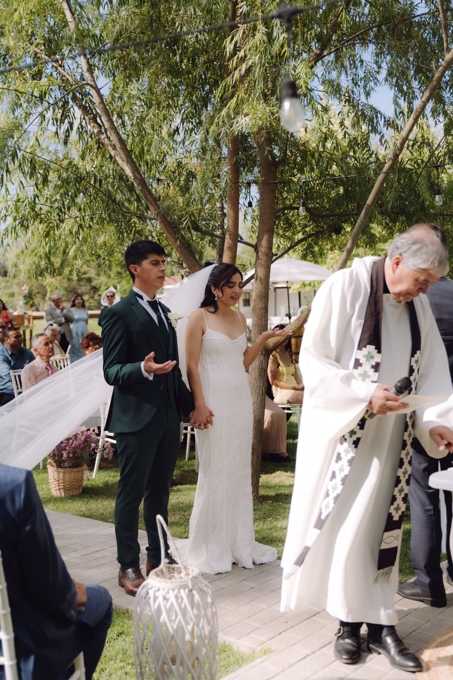 Bride and groom hold hands as officiant in white robes leads outdoor garden wedding ceremony.