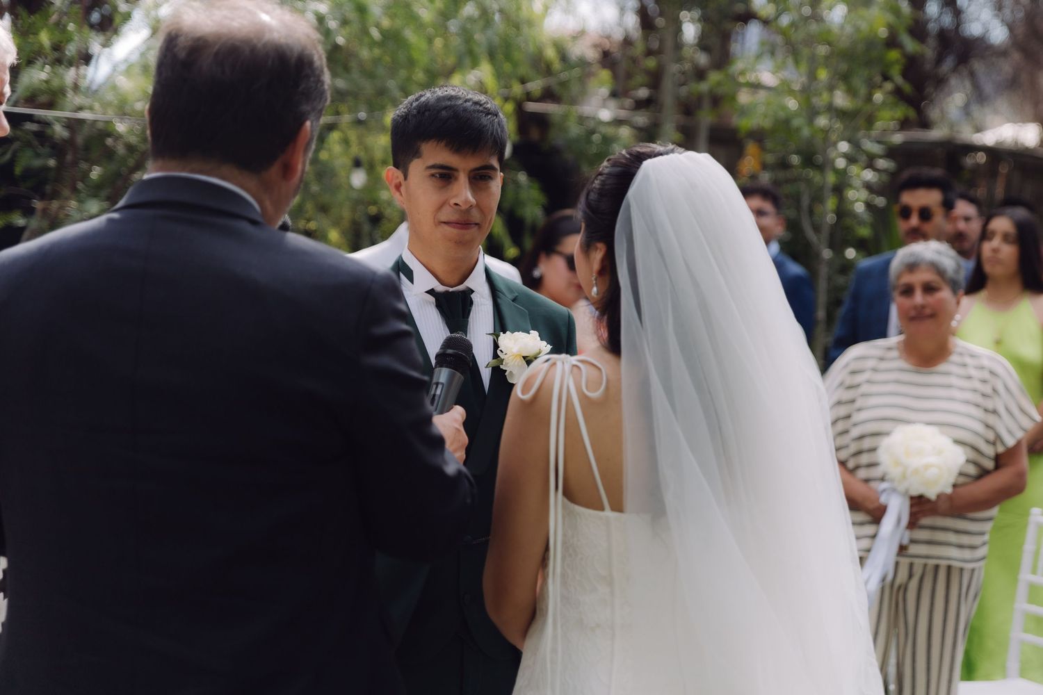 Groom in green suit exchanges vows with bride in white gown at outdoor wedding ceremony.