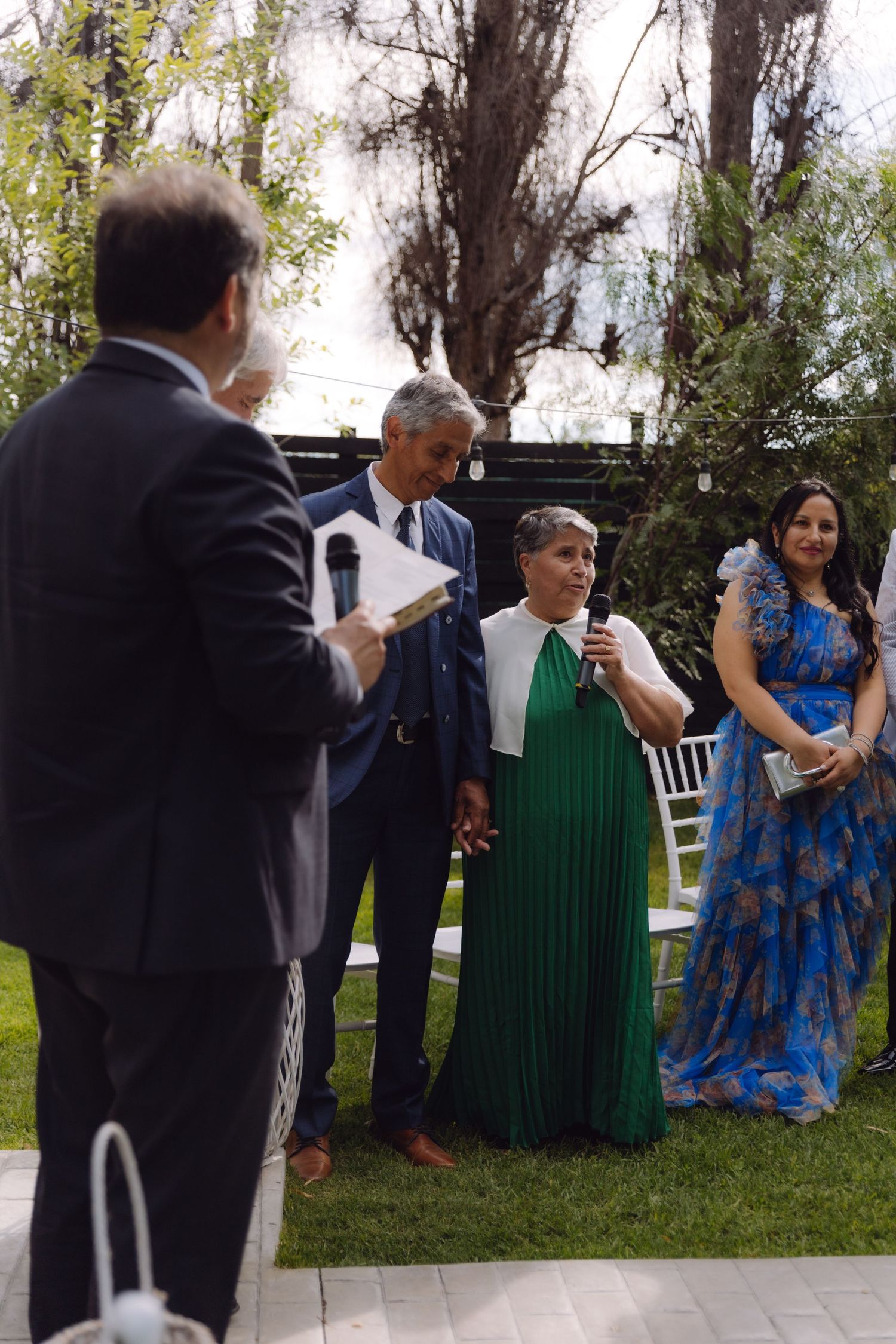 Wedding guests in formal attire speak at microphone during outdoor garden wedding ceremony.