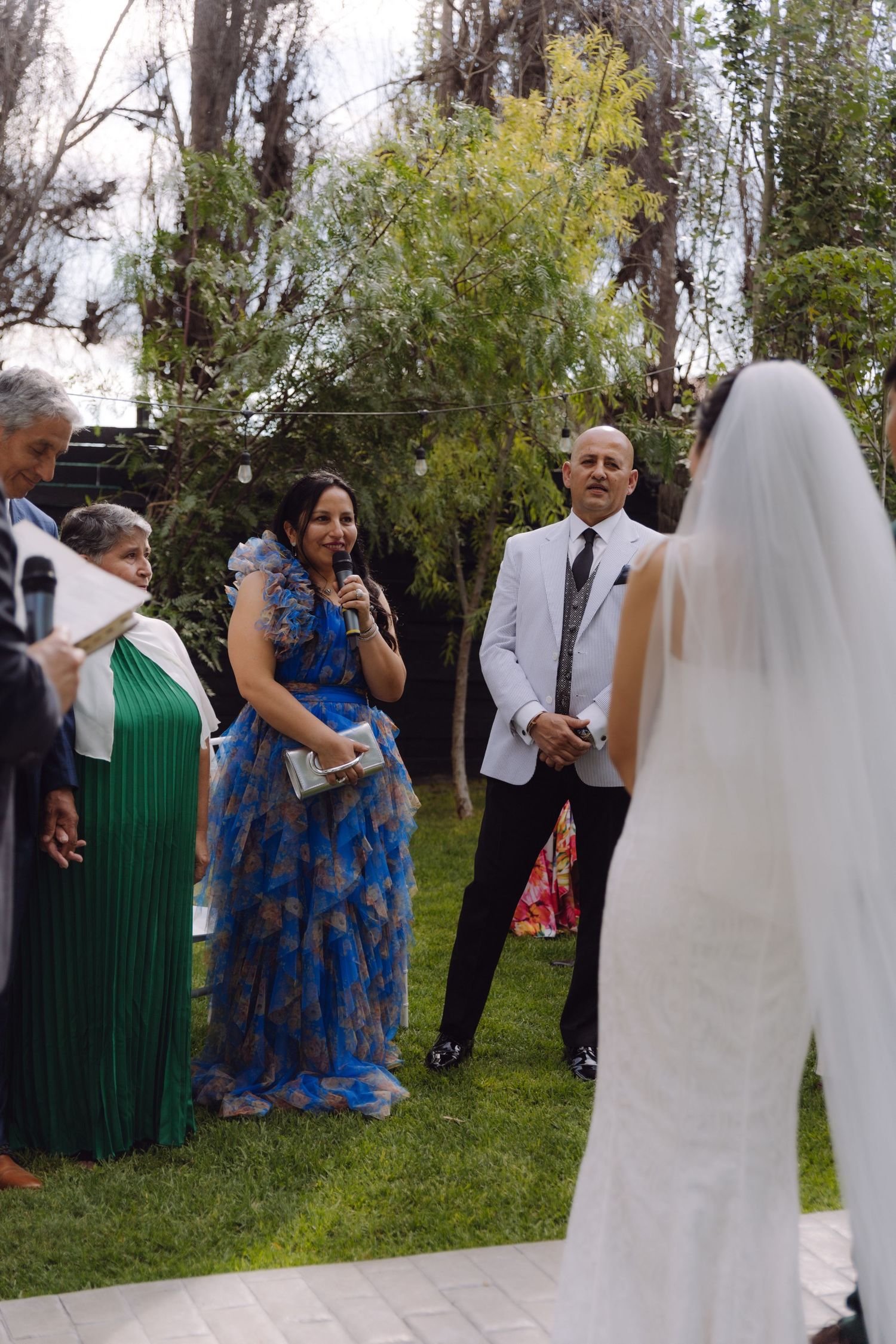 Wedding guest in blue floral dress speaks at microphone during outdoor garden ceremony.