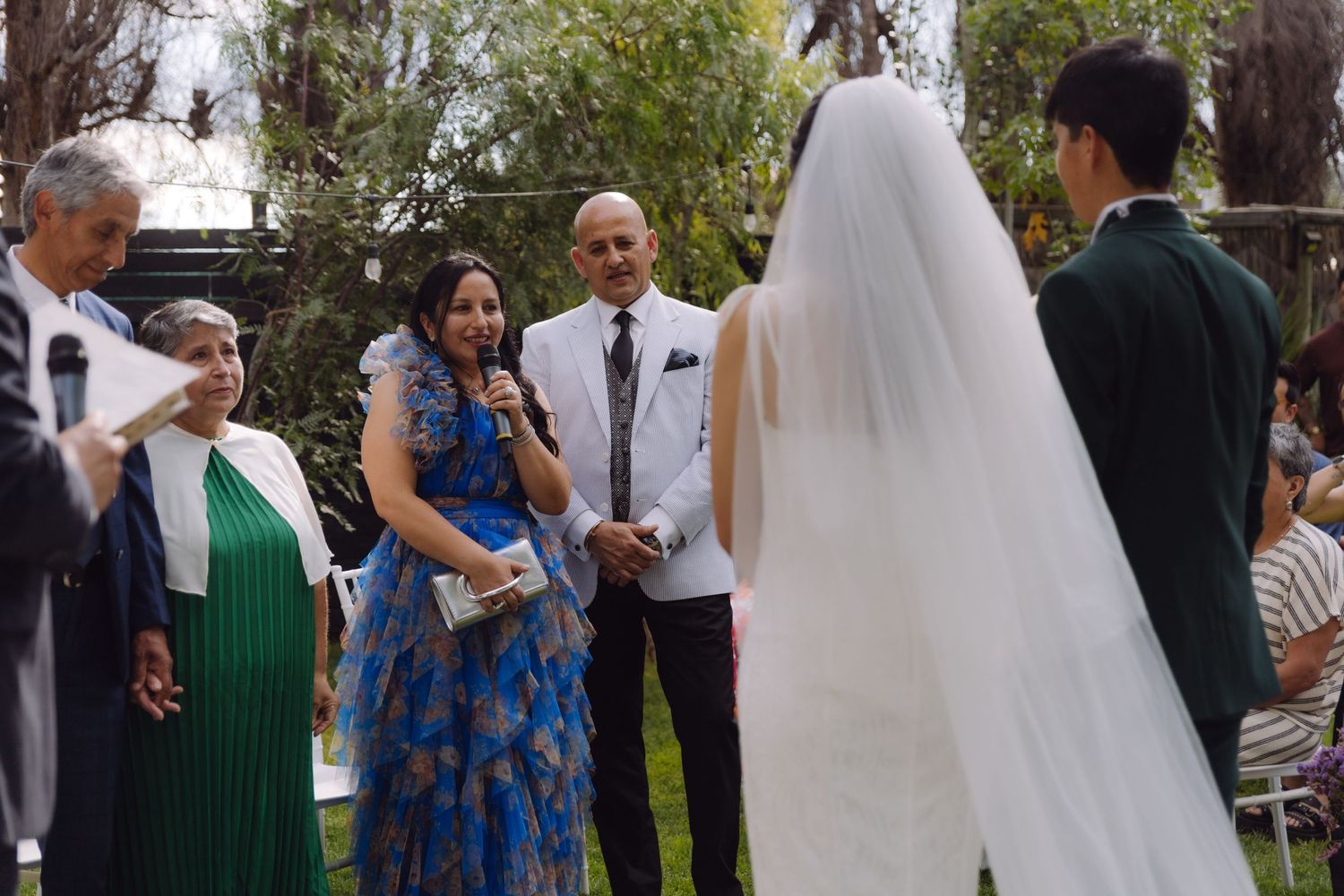 Family members smile as bride and groom exchange vows at outdoor garden wedding ceremony.