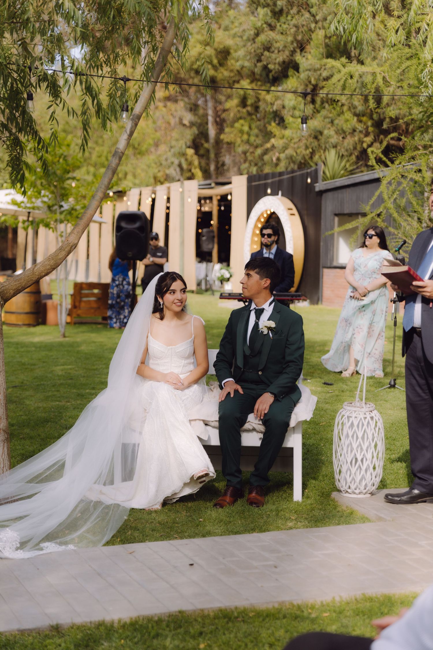 Bride in white gown and groom in green suit sit together during outdoor wedding ceremony.