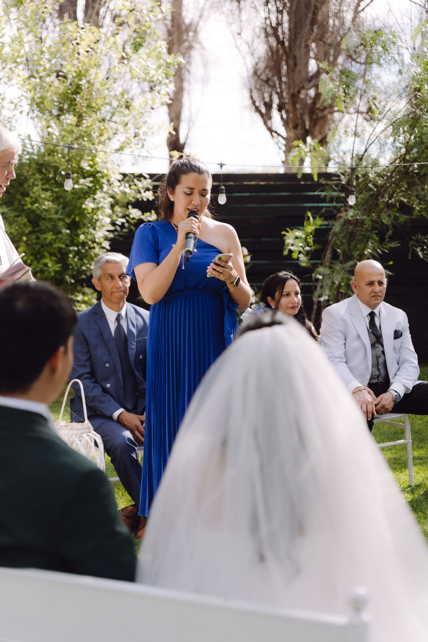 Woman in blue dress speaks at outdoor wedding ceremony into microphone while bride and groom sit nearby.