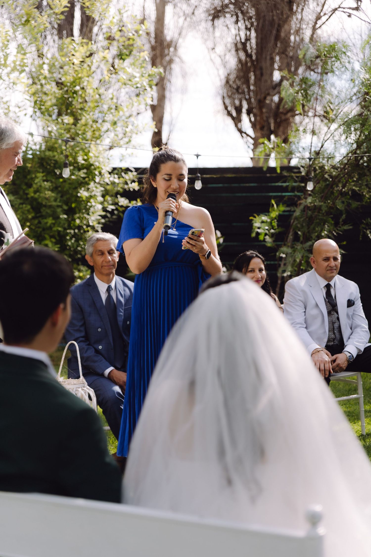 Bridesmaid in royal blue pleated dress reads speech at sunny outdoor wedding ceremony.