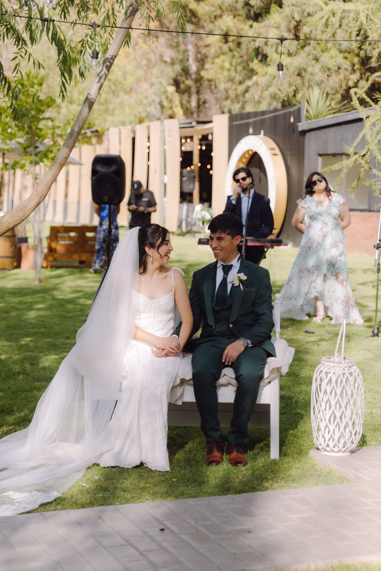 Smiling bride in white gown and groom in green suit sit together during outdoor garden wedding ceremony.