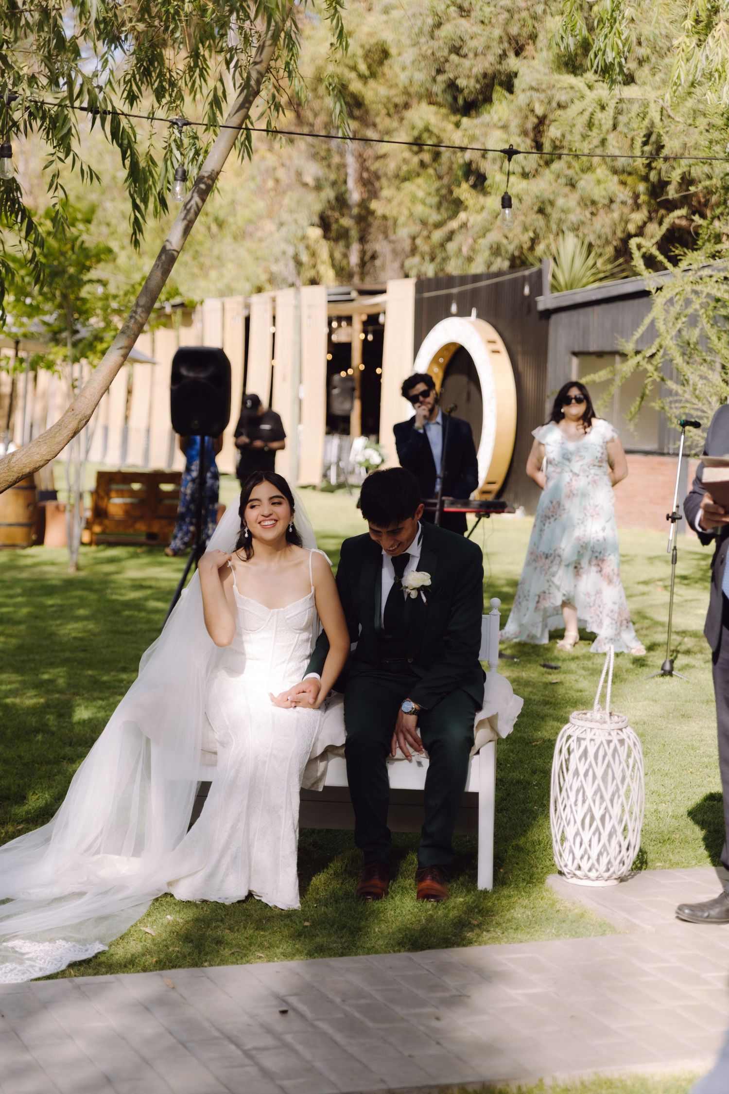 Bride in white dress and groom in dark green suit hold hands during outdoor wedding ceremony on green lawn.