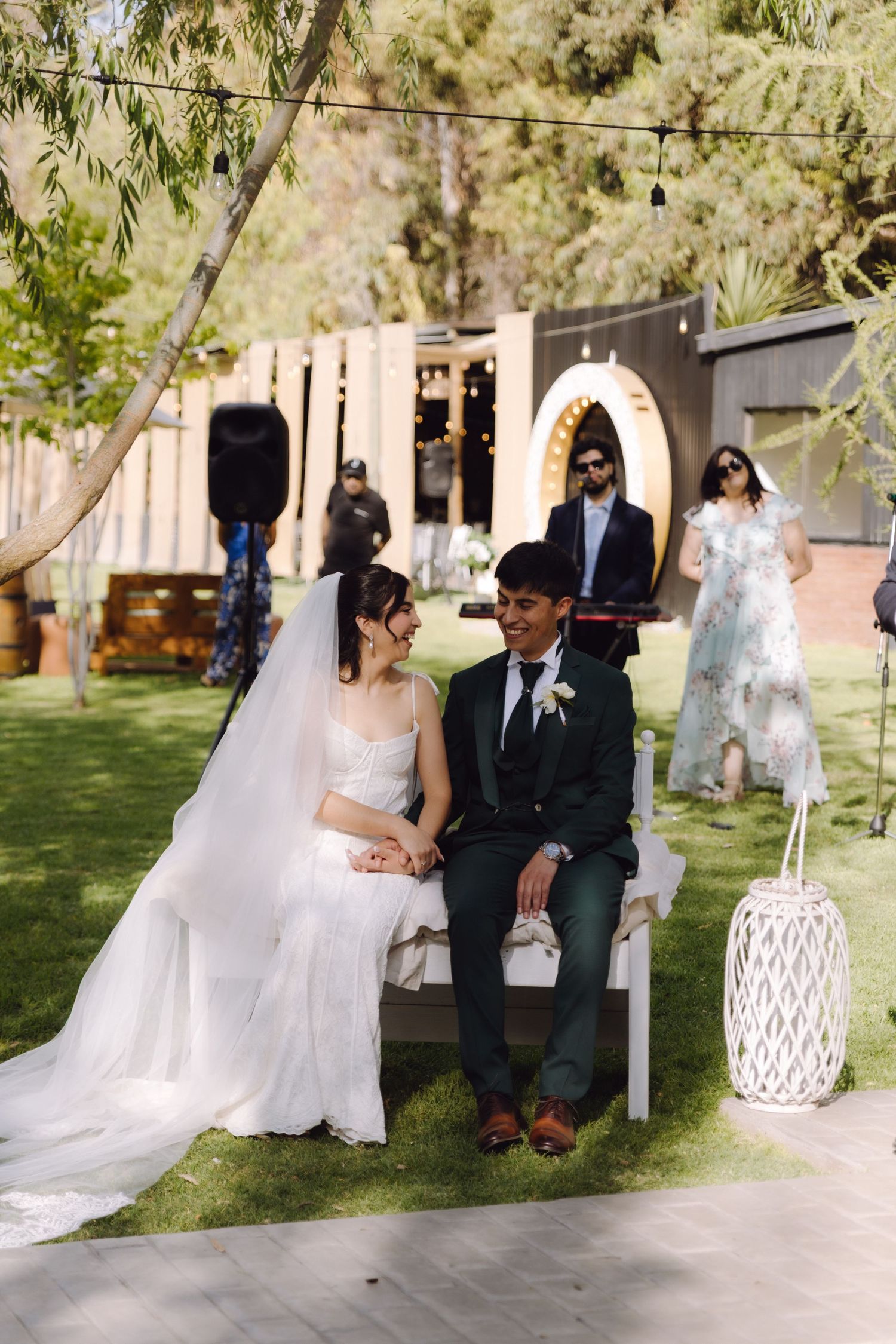 Bride and groom sit together smiling during outdoor garden wedding ceremony with string lights overhead.