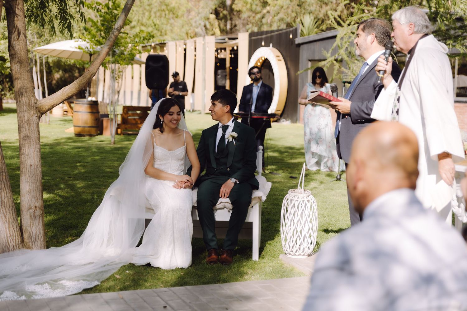Bride and groom seated at outdoor wedding ceremony as officiant reads vows in sunlit garden setting.