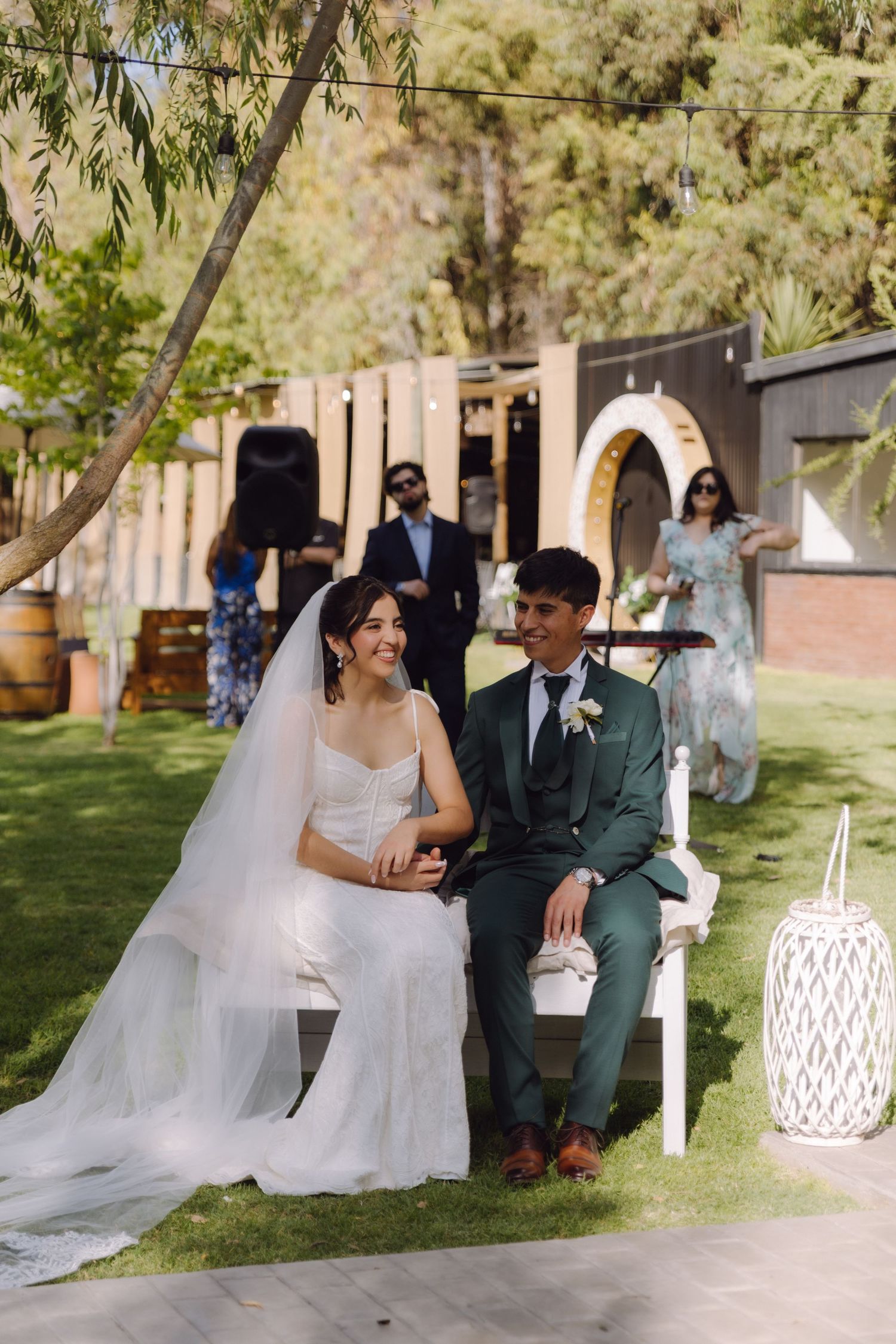 Smiling bride and groom sit together on white bench during sunny outdoor garden wedding.