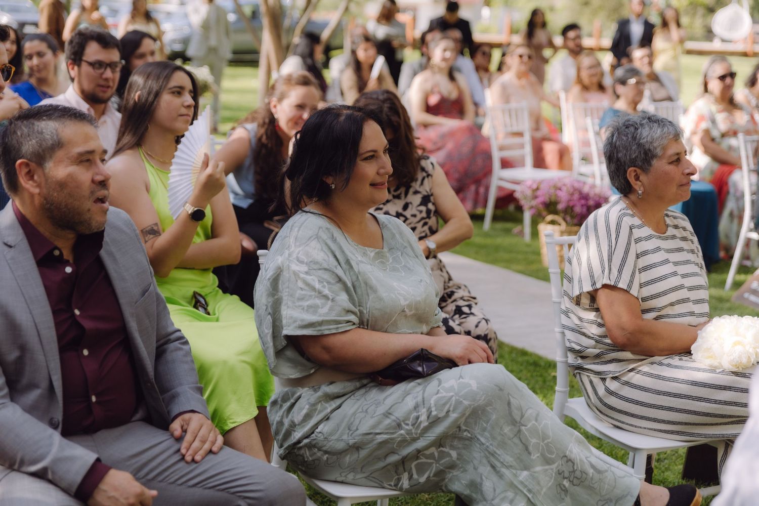 Wedding guests laugh and smile while watching outdoor garden wedding ceremony on a sunny day.