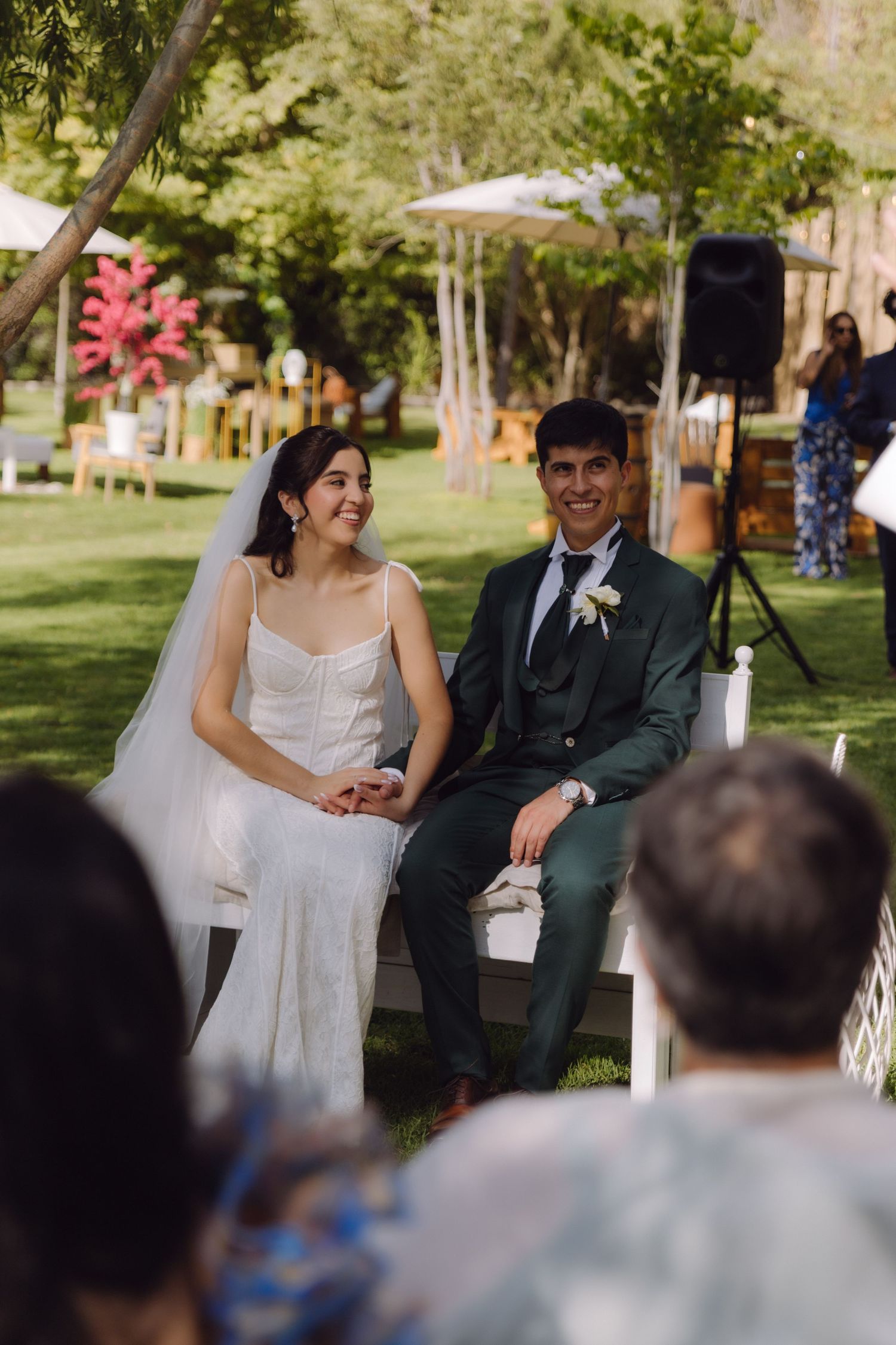 Smiling bride in white gown and groom in green suit sit together during outdoor garden wedding.