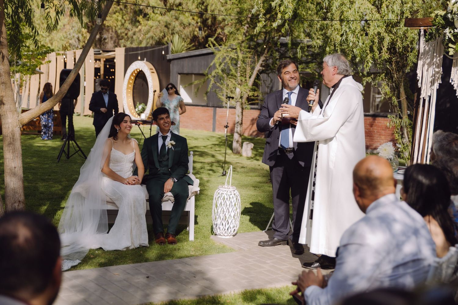 Outdoor wedding ceremony with bride and groom seated as officiant speaks before guests in a garden.