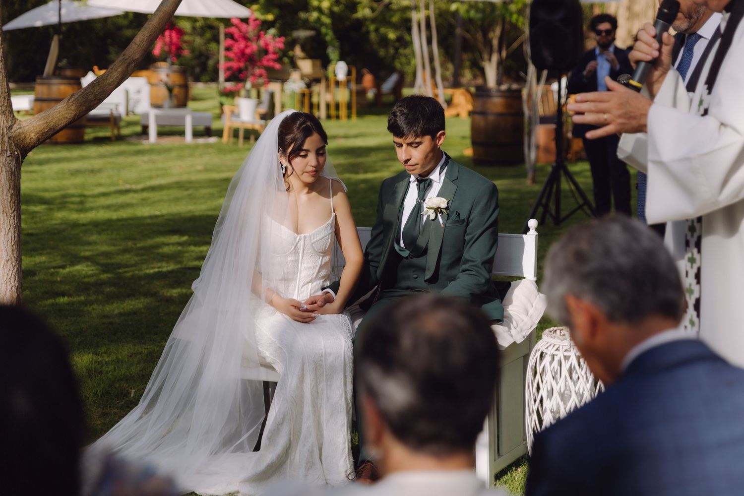 Bride in white gown and groom in green suit sit together during outdoor wedding ceremony.