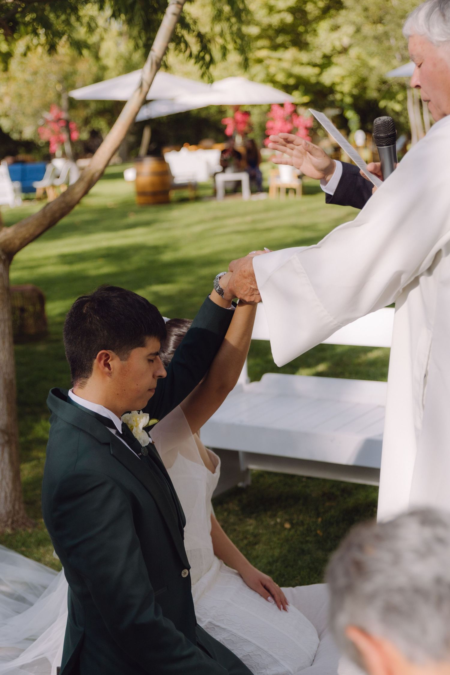 Officiant blesses kneeling bride and groom during intimate outdoor garden wedding ceremony.