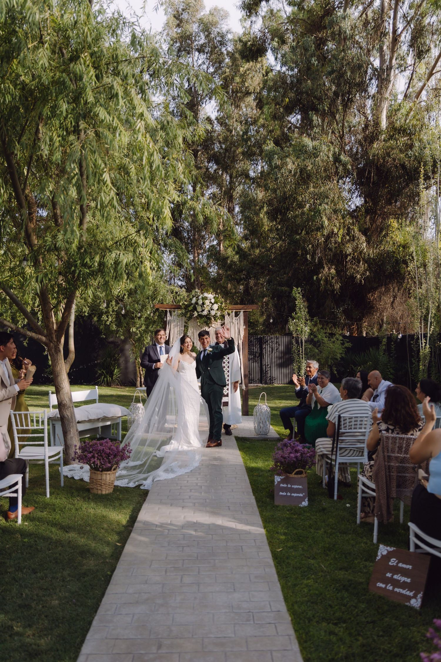 Newlyweds celebrate walking down aisle as guests applaud at outdoor garden wedding ceremony.