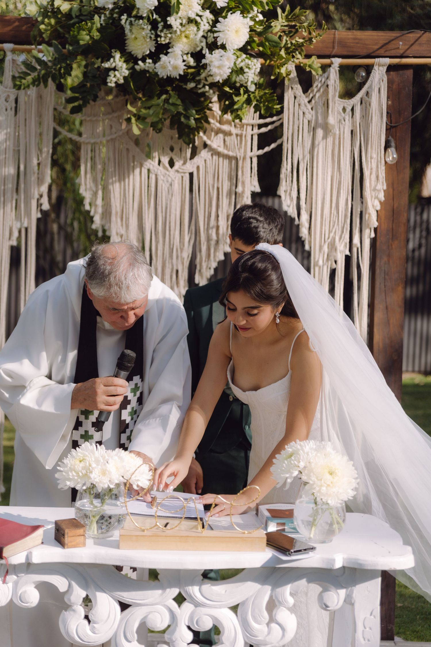 Bride and groom sign wedding documents at decorated altar under macrame boho wedding arch.