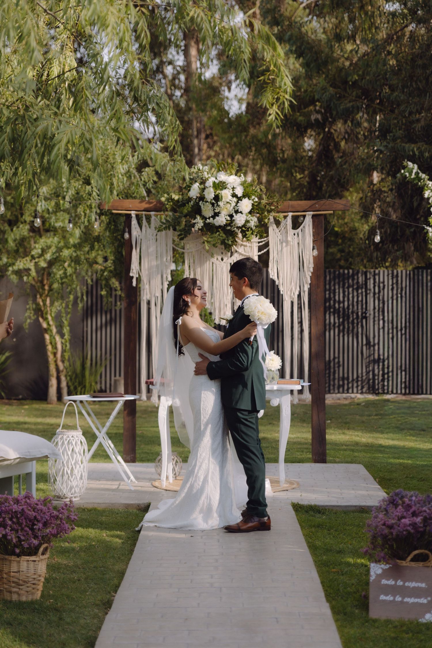 Newlyweds embrace at elegant outdoor wedding ceremony with macrame arch and white flowers.
