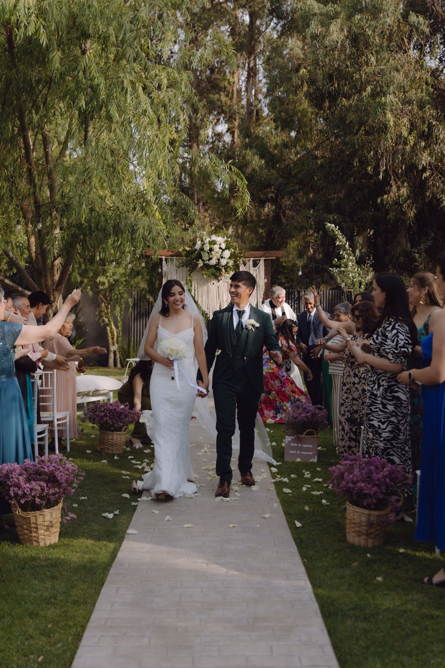 Newlyweds walk down aisle as guests celebrate with flower petals at outdoor wedding.