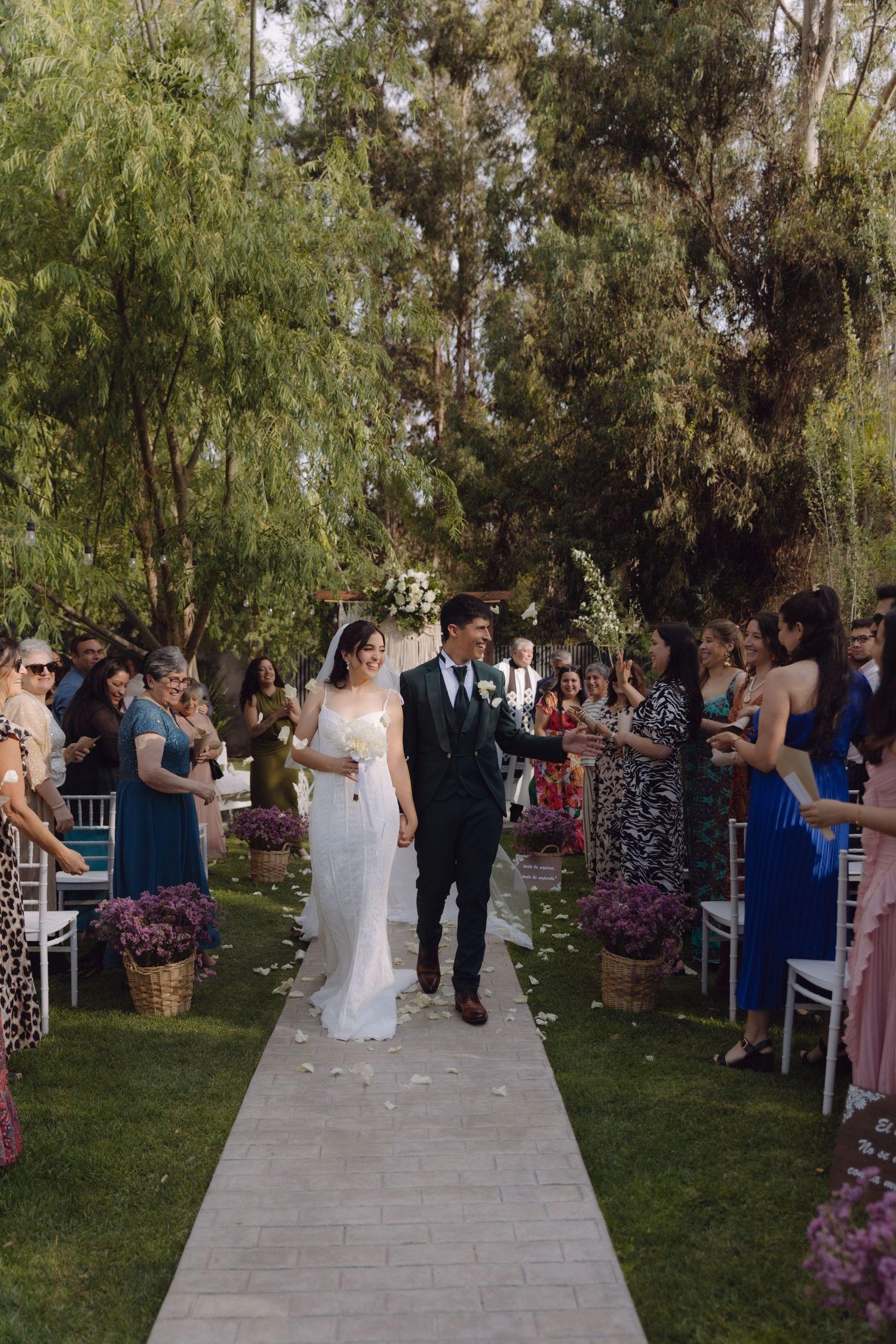 Newlyweds walk hand in hand down petal-lined aisle at lush outdoor garden wedding.