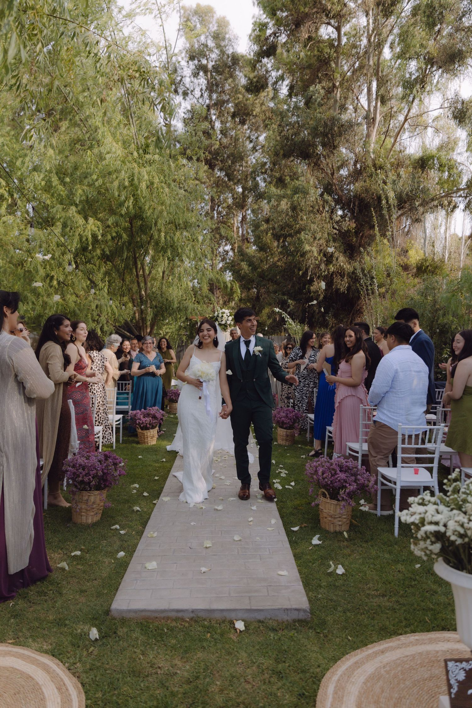 Bride and groom exit ceremony down white aisle lined with purple flowers and guests.