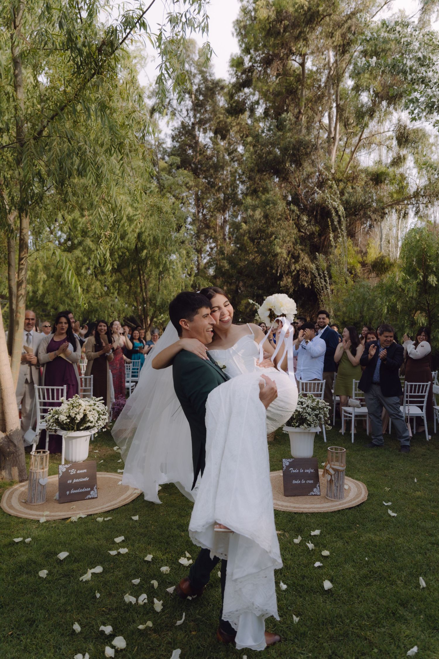 Groom lifts bride in arms celebrating after outdoor garden wedding ceremony with guests.