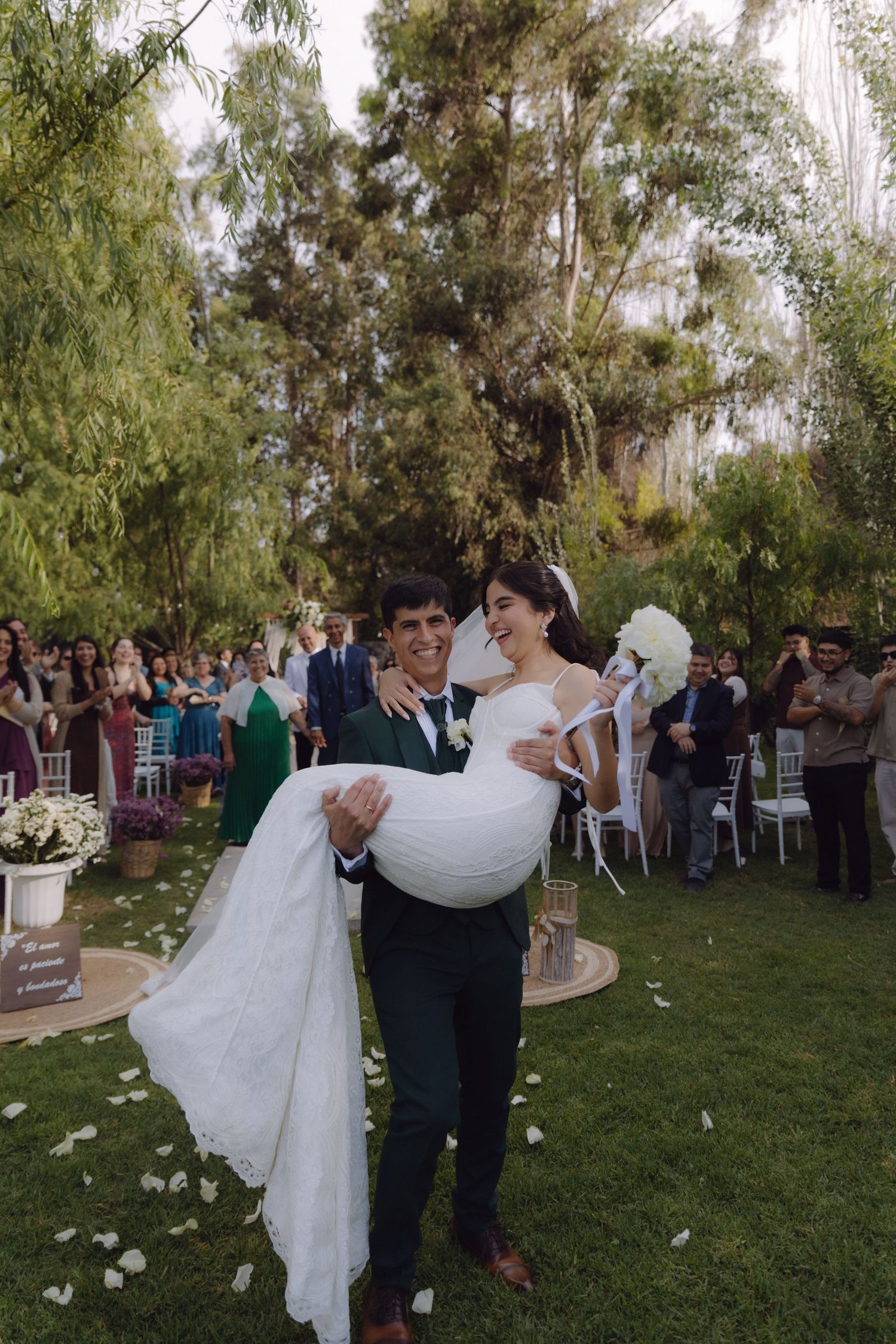 Groom carries bride in white gown after outdoor wedding ceremony with guests cheering.