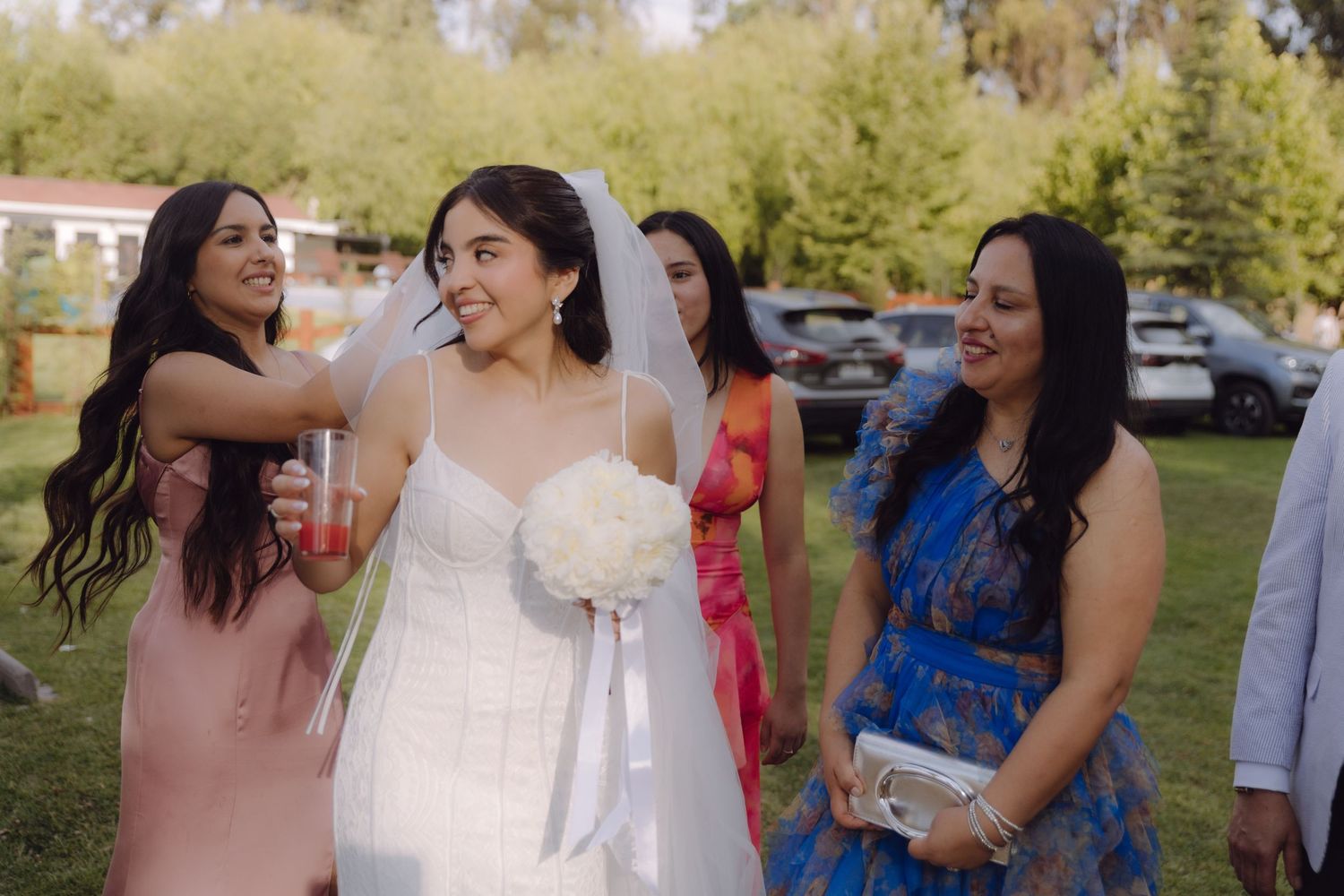 Smiling bride in white gown holds bouquet surrounded by guests in colorful dresses.