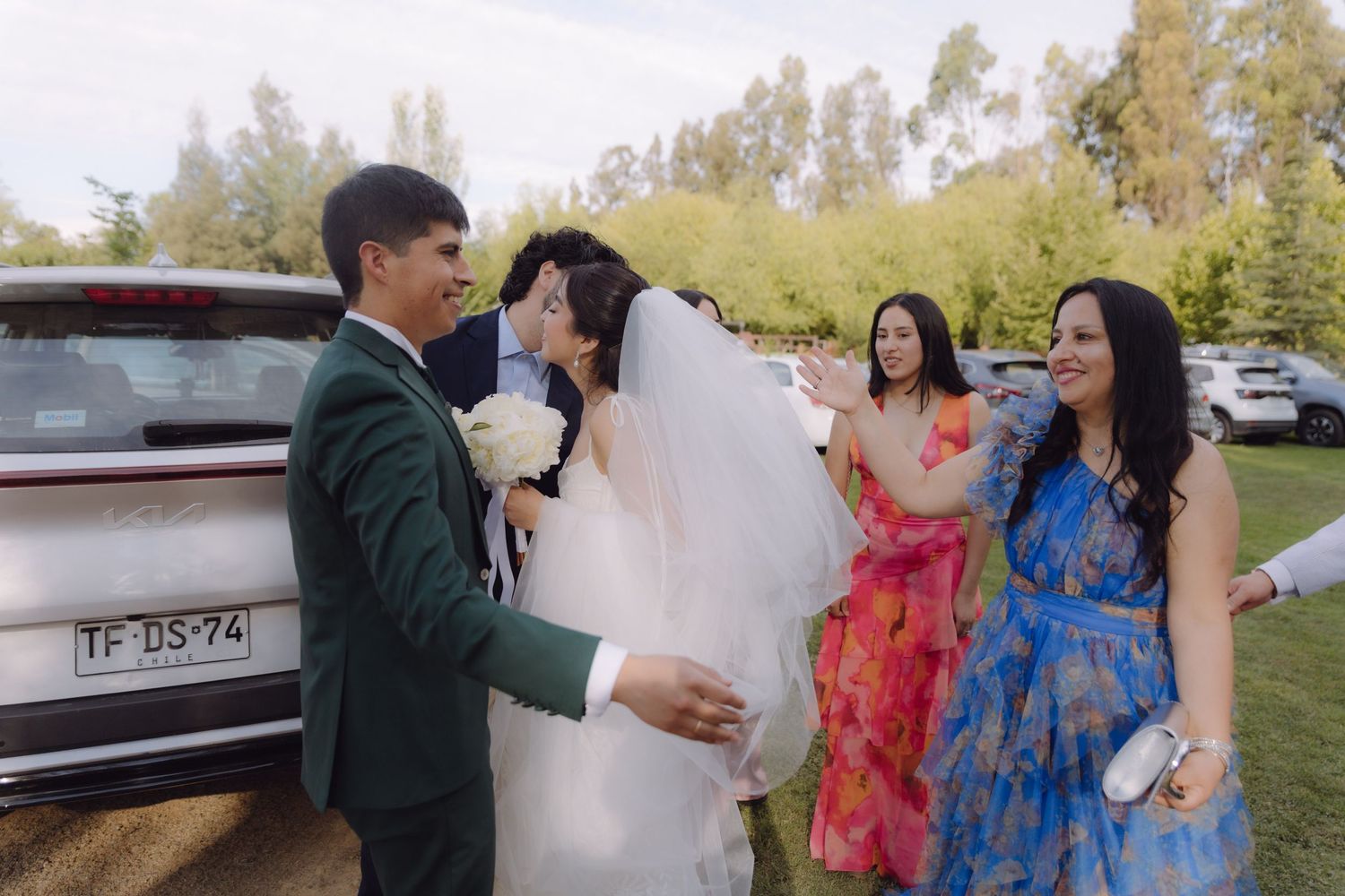 Wedding couple shares kiss near car as guests in colorful dresses celebrate outside.
