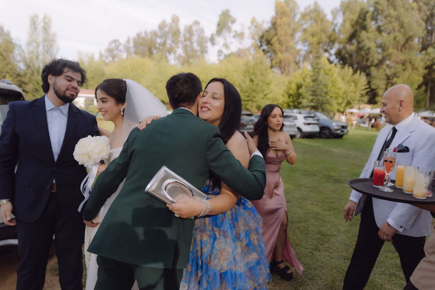 Groom in green suit hugs guest warmly as bride holds bouquet at outdoor wedding.