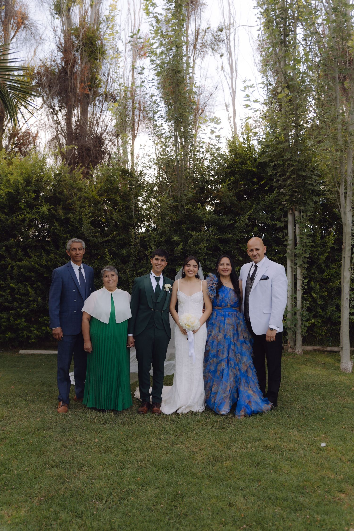 Wedding party group photo with bride, groom, and family in garden setting.