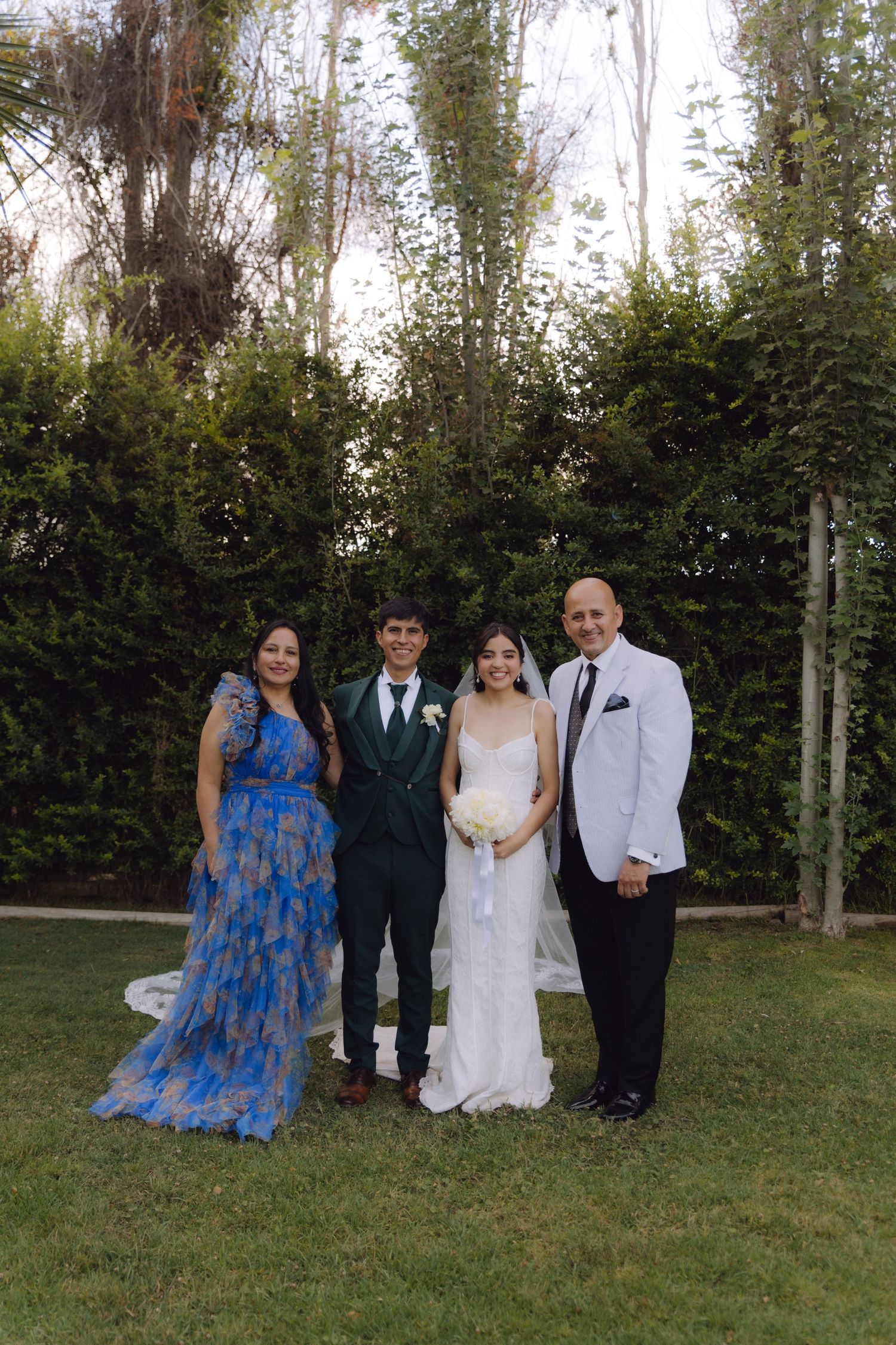 Bride and groom pose with parents in blue and white formal attire outdoors.
