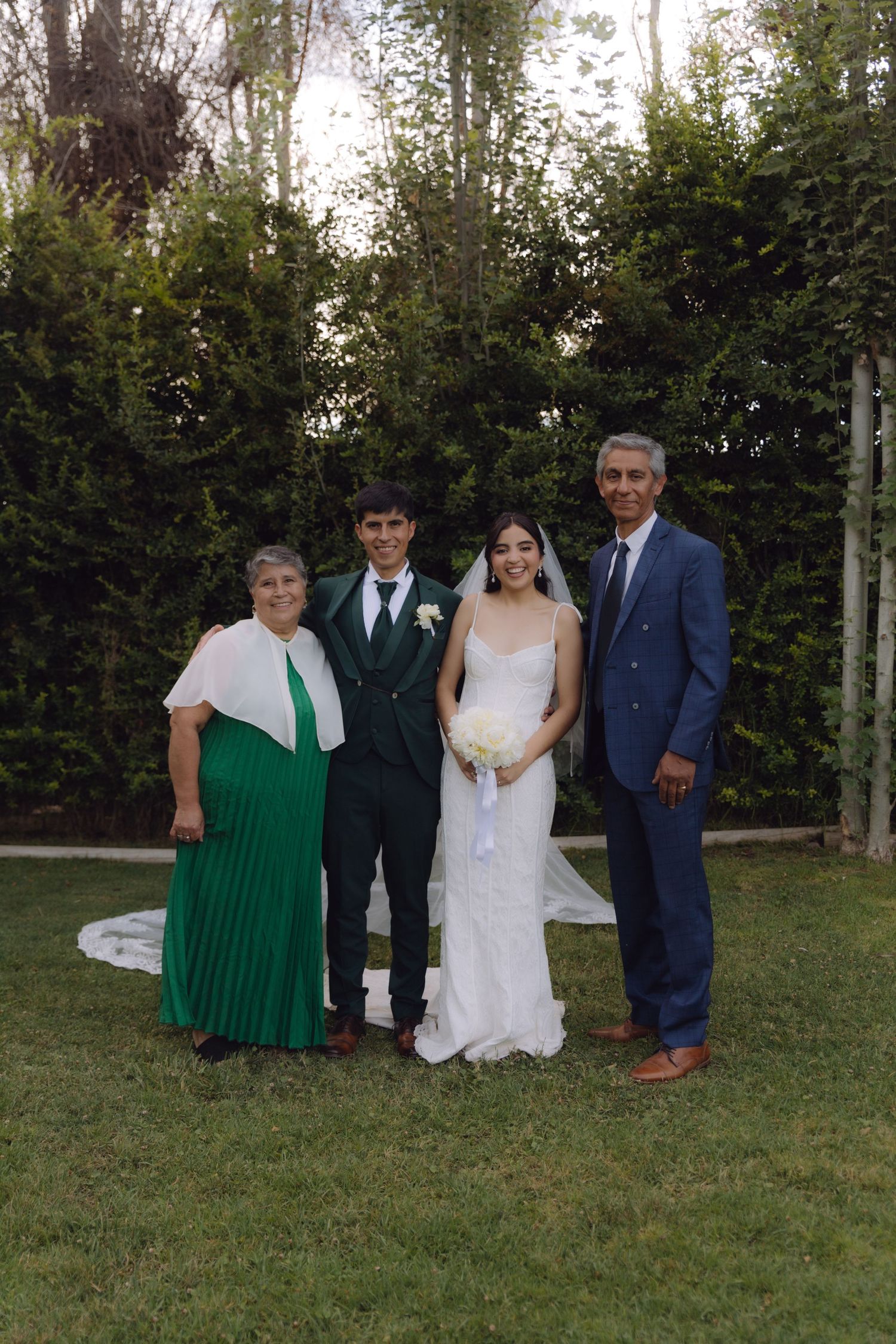 Bride and groom smile with parents in green dress and blue suit in garden.