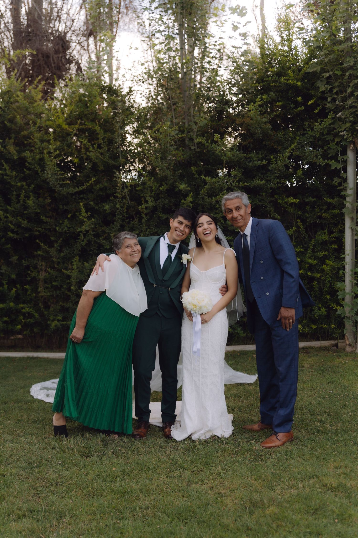 Bride and groom pose warmly with parents in lush green garden setting.