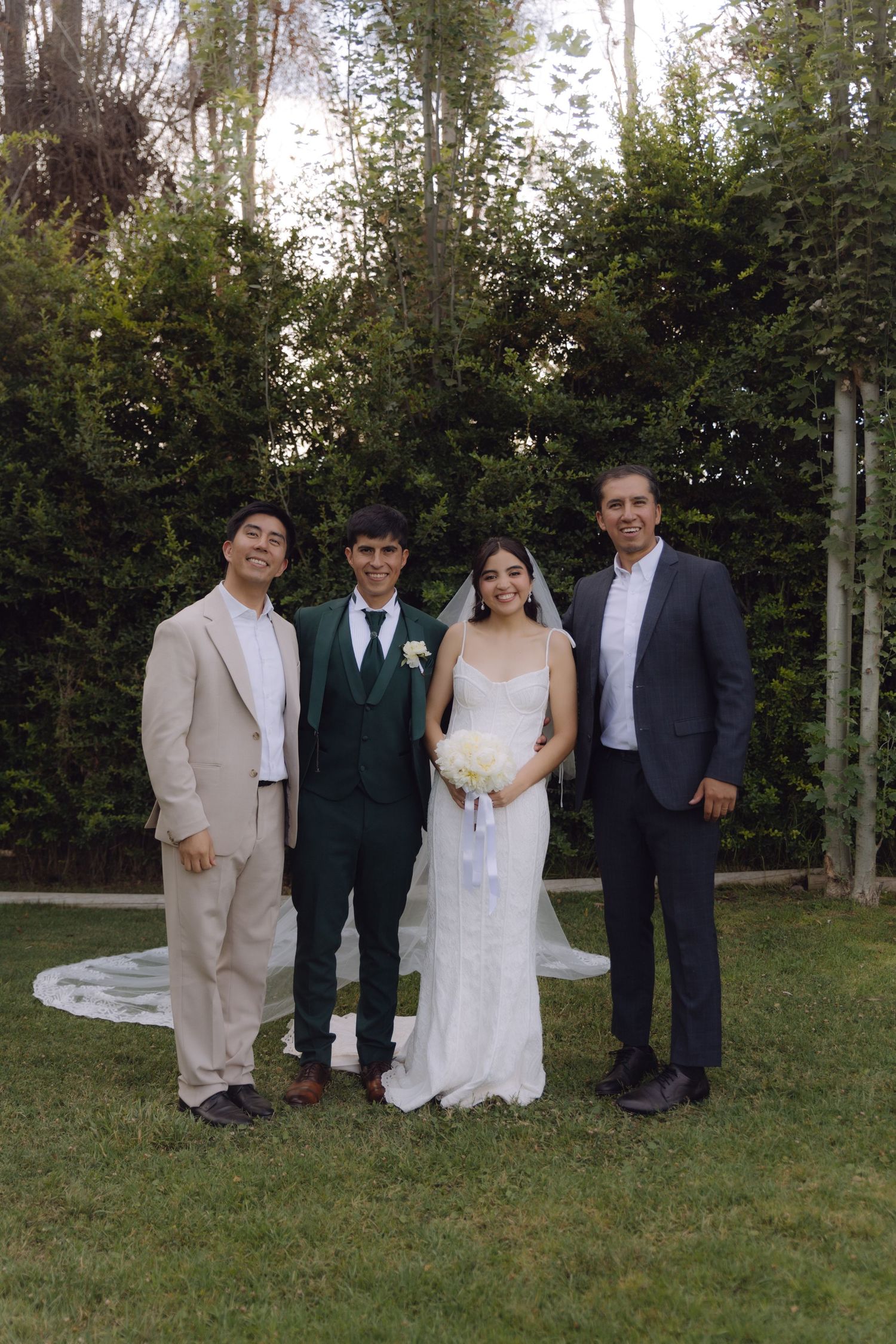 Bride and groom pose with two guests in a lush garden setting during an outdoor wedding celebration.