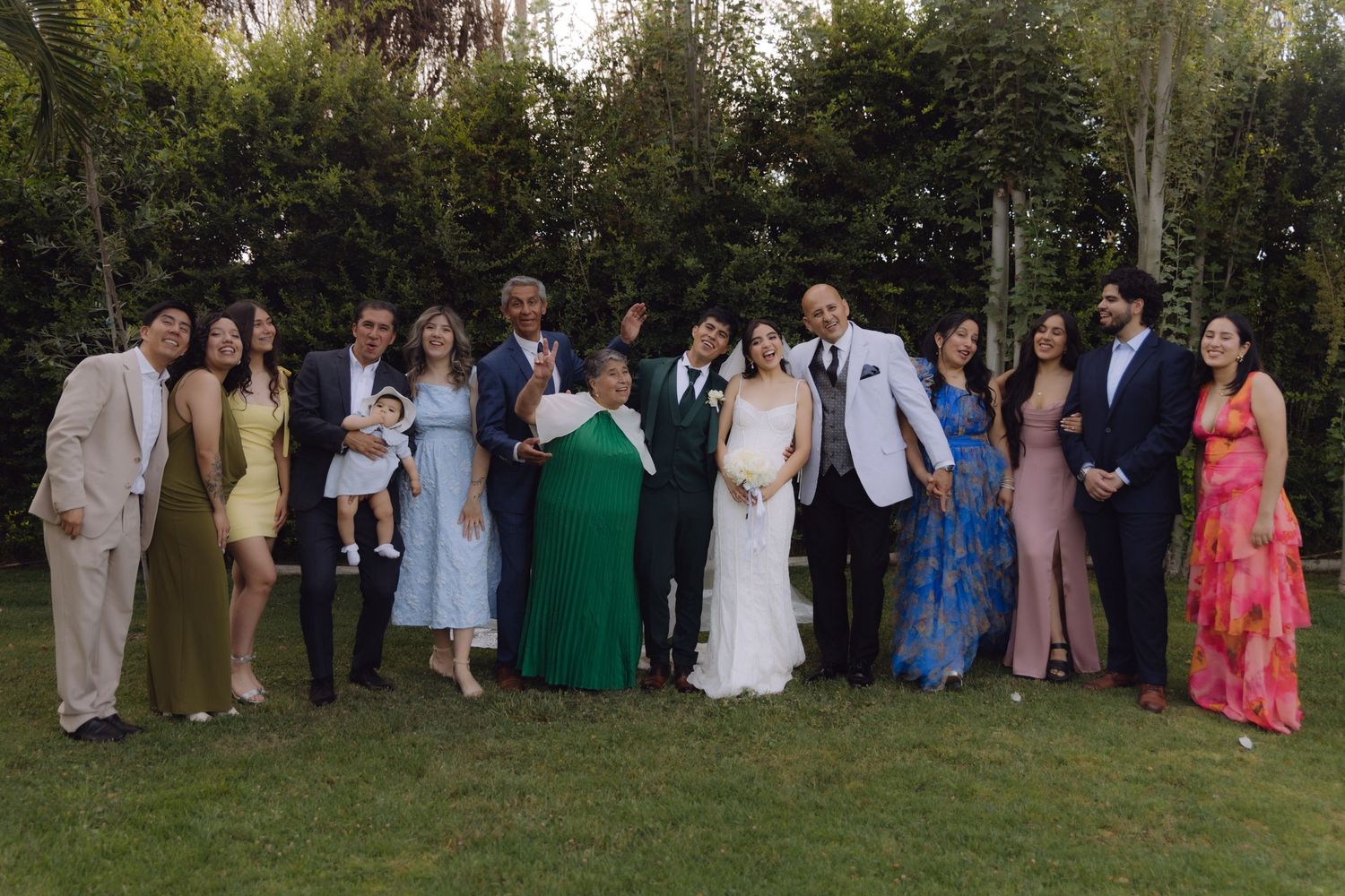 Large wedding family group portrait taken outdoors on green grass with tall hedges in the background.