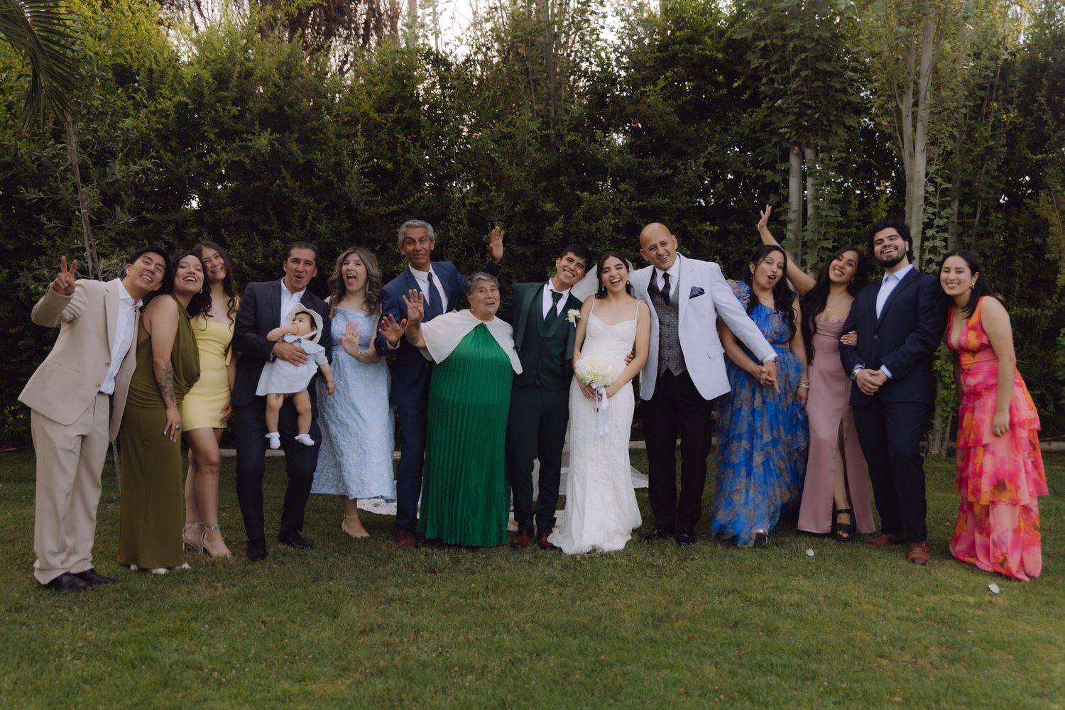 Joyful wedding party group photo with guests posing and celebrating on a garden lawn.