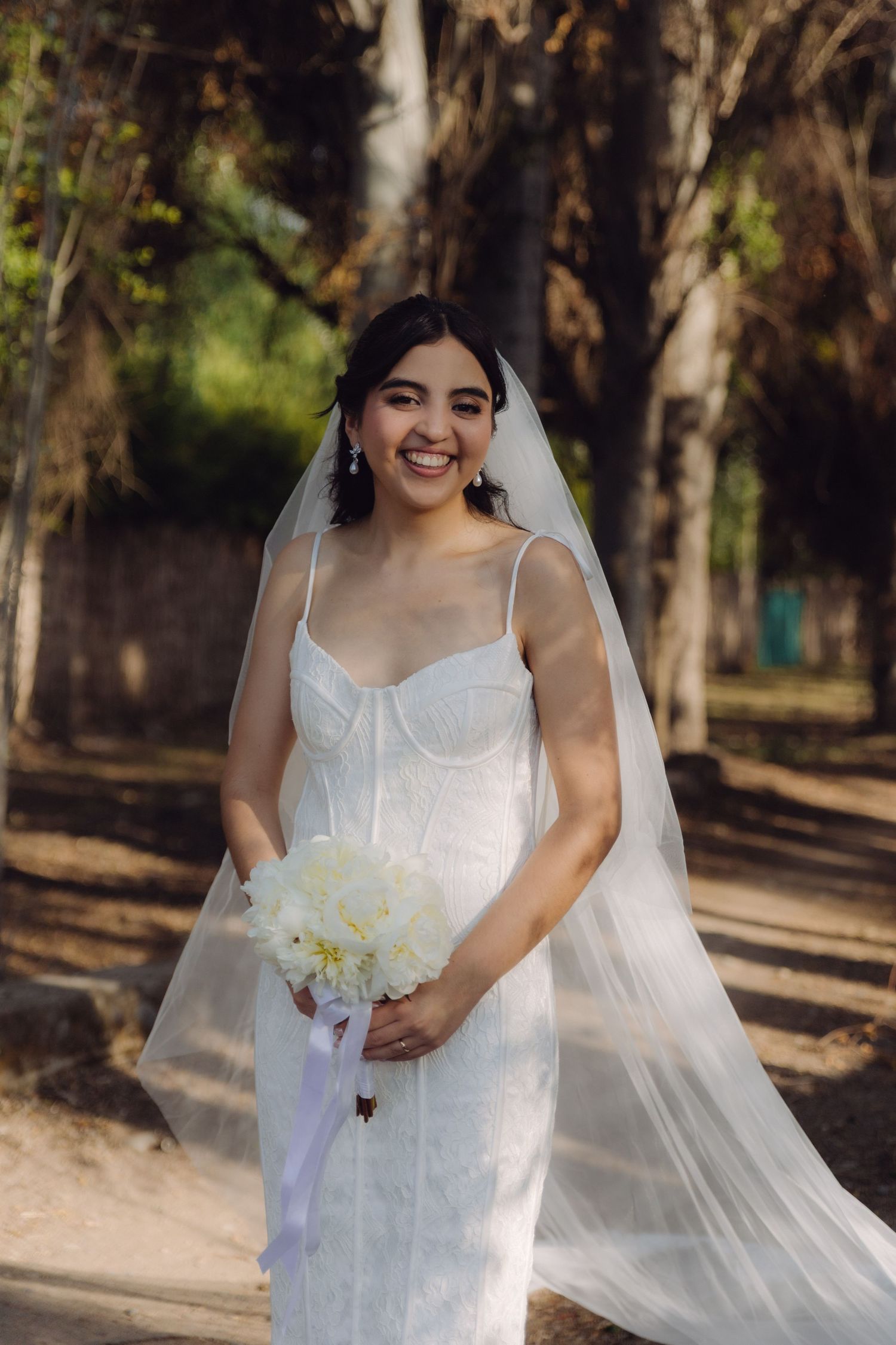 Smiling bride in white lace gown and veil holds white bouquet in a sunlit outdoor setting.